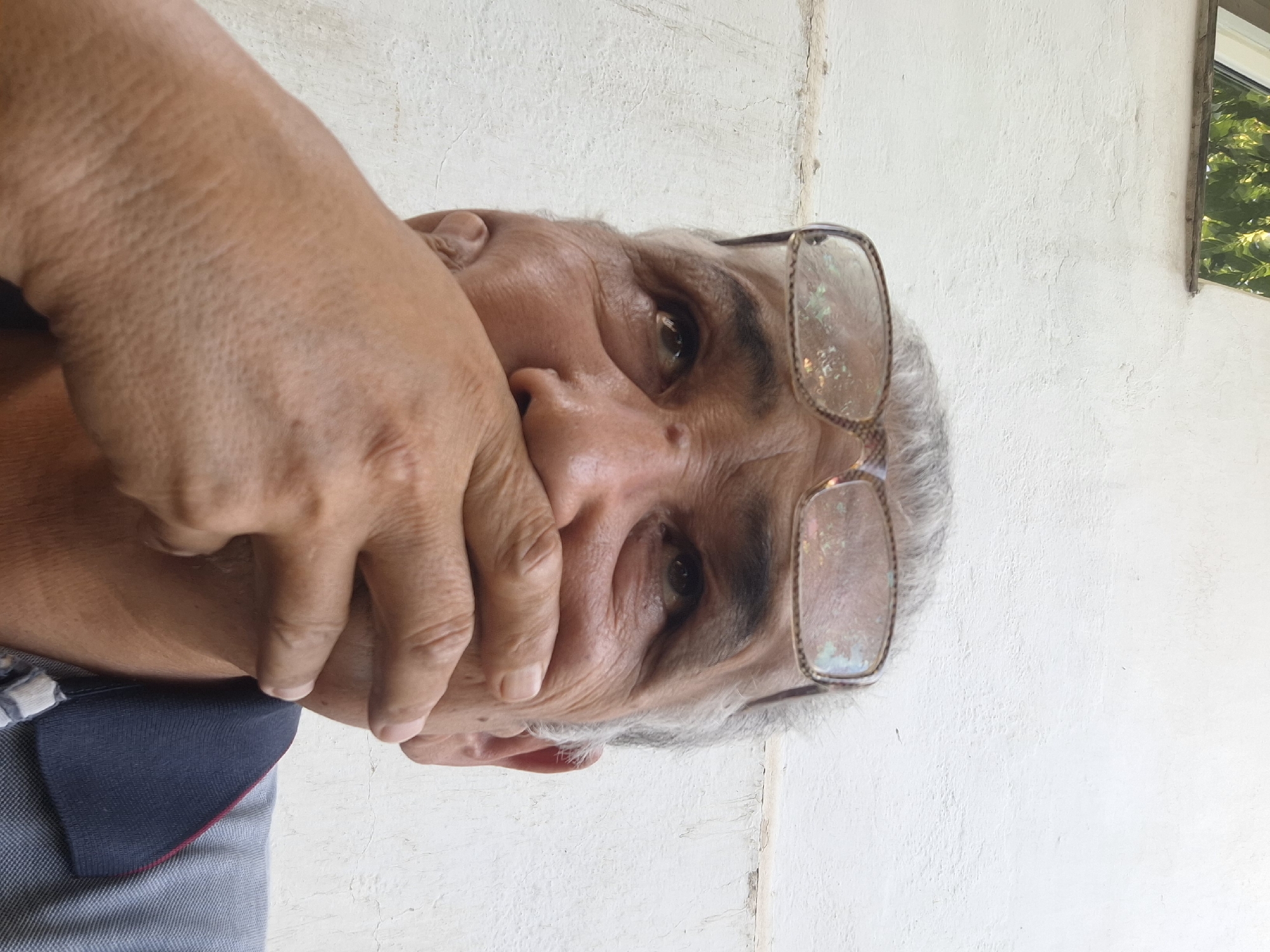 The image shows a close-up, low-angle shot of an elderly man's face, partially obscured by his hand. He is wearing glasses with a light-colored frame, which appear to have some smudges or reflections on the lenses. His hair is gray and sparse. The man's mouth is covered by his right hand, and his eyes are looking upwards and to his right. His skin shows signs of aging, with wrinkles and some texture. He is wearing a blue collared shirt with a red and white trim. The background is a textured white wall, with a small portion of a window visible at the top left, showing green foliage. The lighting suggests an outdoor setting, likely during the day. This scene appears to be located in Almaty, Kazakhstan, based on the available information.