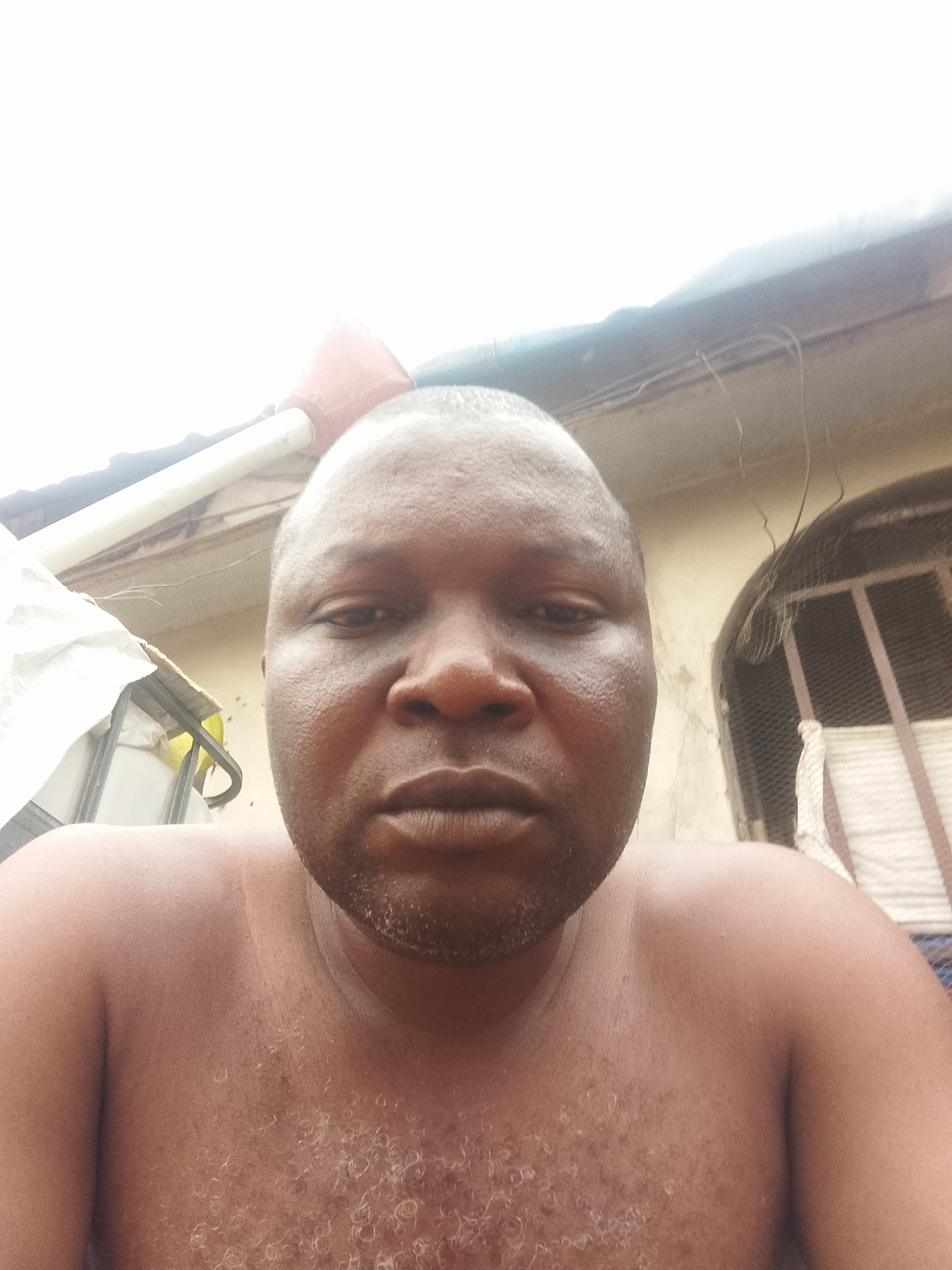 A close-up, high-angle selfie of a dark-skinned, bald man is set against an outdoor backdrop. The man has a coarse beard and appears sweaty. Behind him, there is a building with a corrugated metal window covering on the right. A water tank sits to the left, and a red plunger is visible above the man's head. The location is the city of Unknown, Unknown.