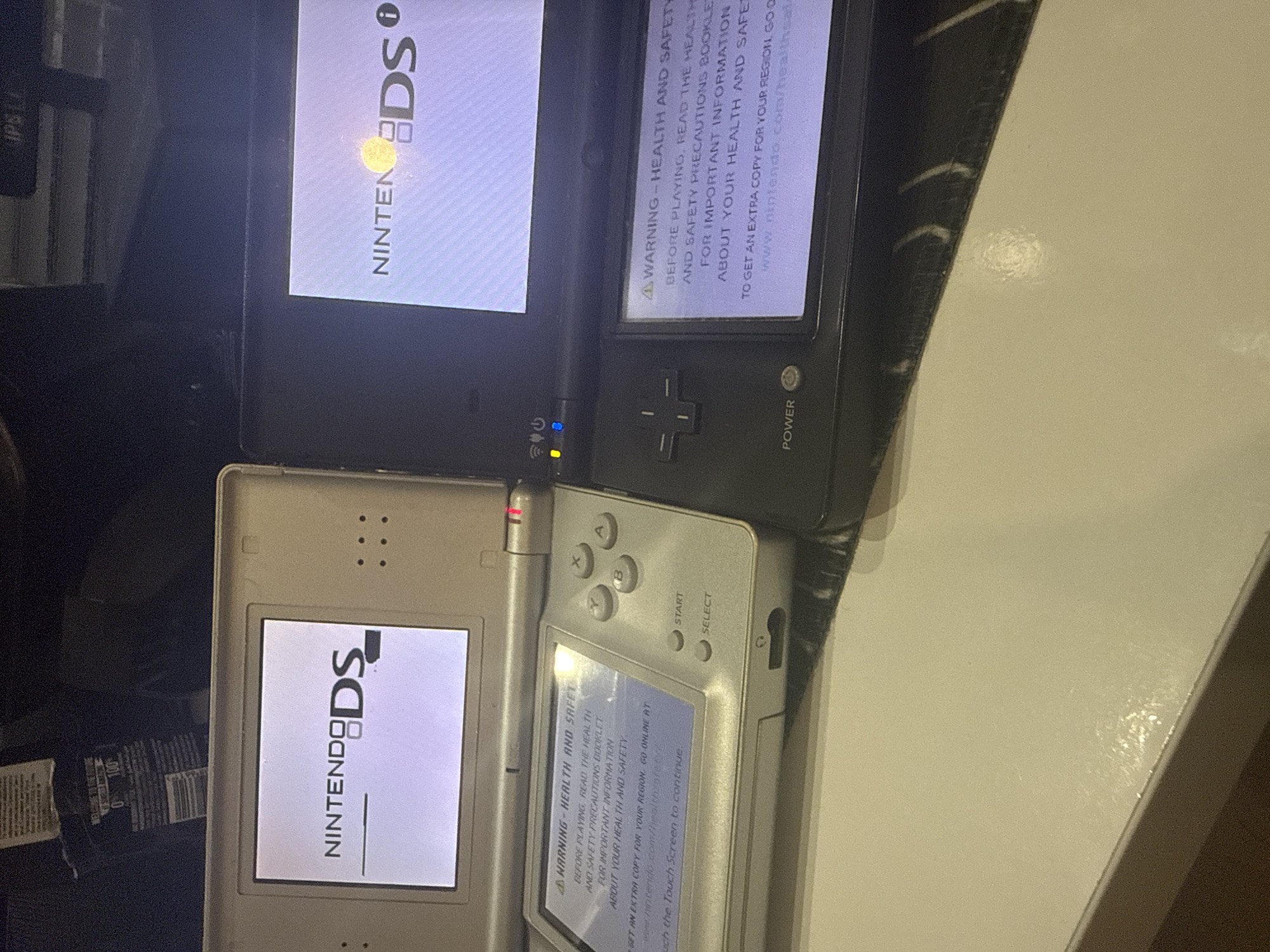 The image shows two Nintendo DS gaming consoles placed on a light-colored surface, likely a table. The console on the left is silver, and the one on the right is black. Both consoles display warning messages on their lower screens regarding health and safety precautions, advising users to read the booklet and visit www.nintendo.com/healthsafety. The upper screen of the silver DS shows the Nintendo DS logo, and the upper screen of the black DS also shows the Nintendo DS logo with a bright yellow light emanating from it. The lighting in the scene is dim, with the screens of the consoles being the primary light sources. The environment appears to be indoors, with some blurry objects in the background. There are no people visible in the image. The location is not specified in the image or the provided crops.