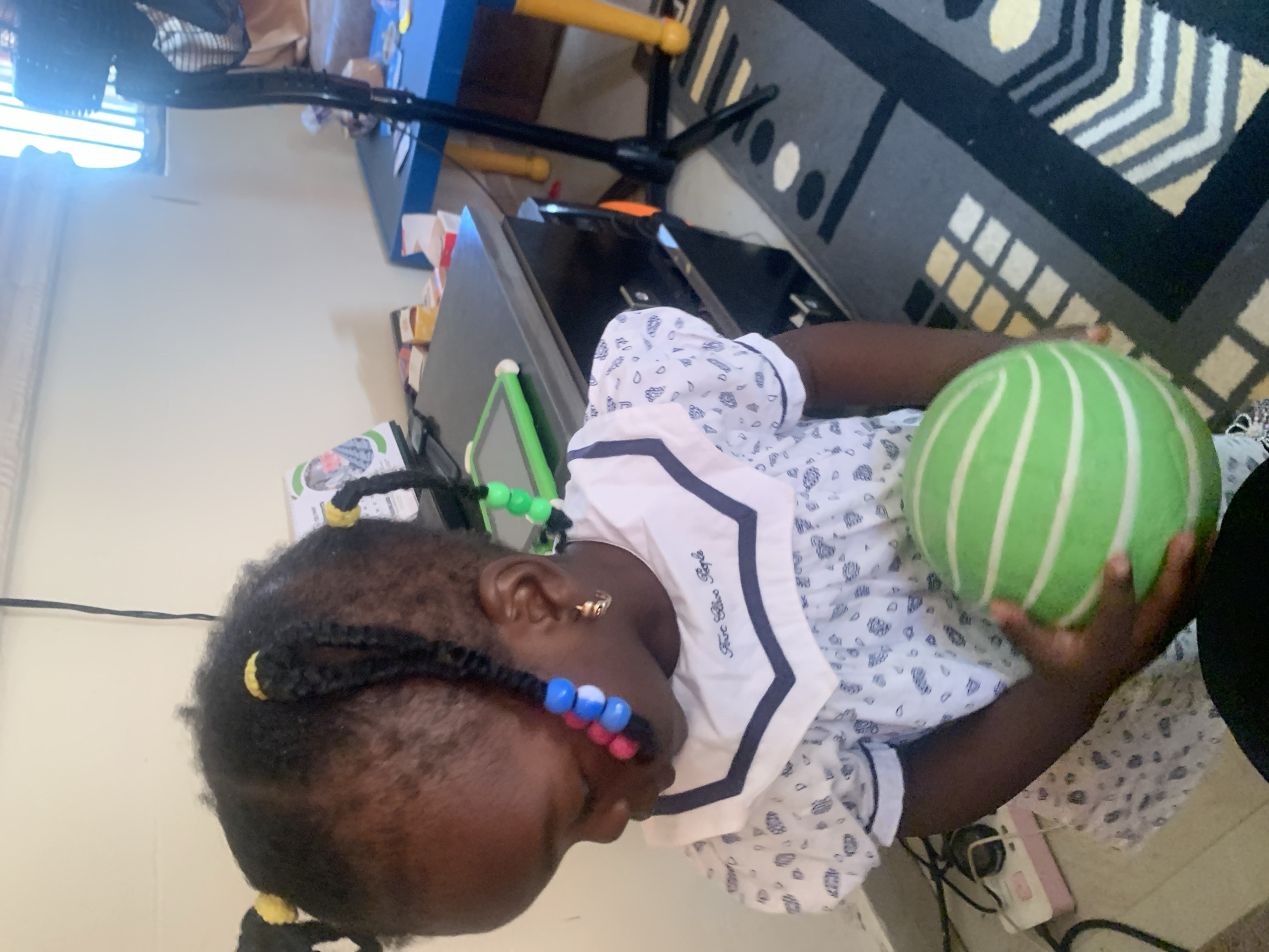 The image shows a young Black girl with braided hair adorned with colorful beads. She is wearing a white dress with a blue collar and a patterned design. The girl is holding a green and white striped ball in her hands. The background appears to be an indoor setting, possibly a room in a home or a school in Gura Top, Nigeria. Various objects are visible in the background, including furniture, a tablet, and a fan. The lighting suggests it is daytime, with some natural light coming from a window.