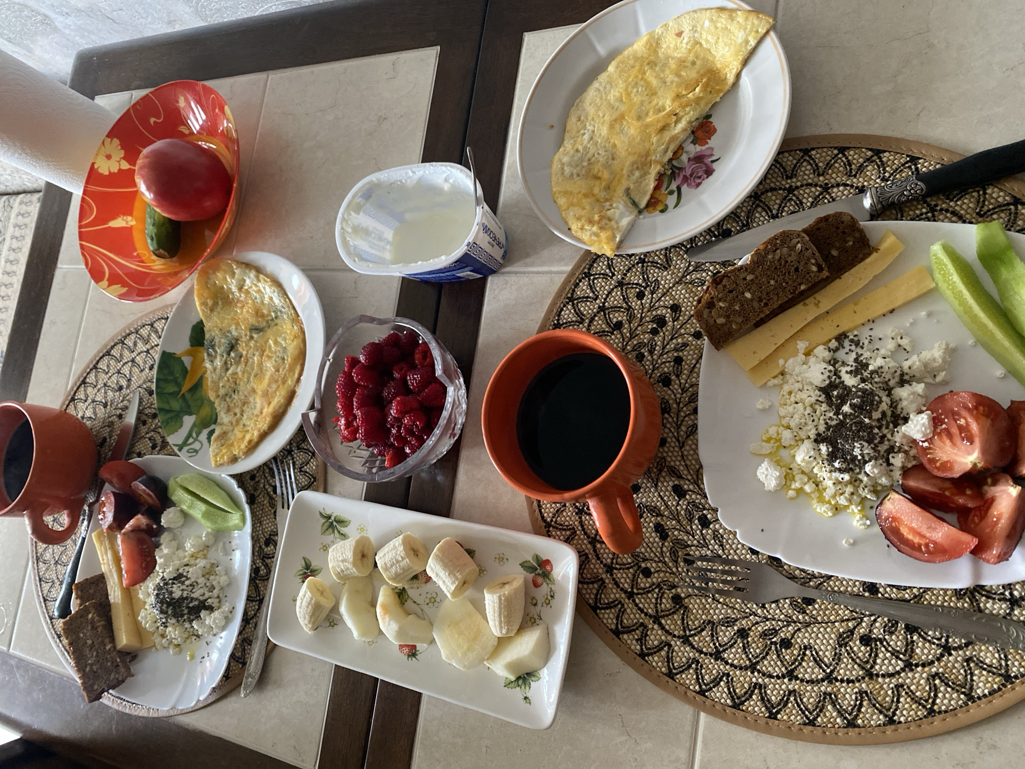 A top-down, eye-level shot captures a rustic breakfast spread laid out on a tiled surface, possibly a table. The scene is rich with various food items arranged on plates and in bowls. In the foreground, a white plate holds a generous portion of cottage cheese speckled with chia seeds, accompanied by slices of tomato and cucumber. Beside it, two slices of dark bread and slices of yellow cheese are neatly arranged, with a knife featuring an ornate handle resting on the plate.
To the left, a rectangular white dish showcases sliced bananas and pears, decorated with small red strawberry illustrations. Above this, on a circular woven placemat, sits an orange mug filled with dark coffee, next to a plate containing cottage cheese, tomato slices, cucumber, and two slices of dark bread with cheese.
Further back, a red bowl with a yellow floral pattern holds a whole tomato and a cucumber. Adjacent to it, a glass bowl is filled with raspberries. Another white plate features an omelet. A container of yogurt with a spoon completes the arrangement. The overall impression is a hearty and visually appealing breakfast, suggesting a calm morning meal in Lida, Belarus.