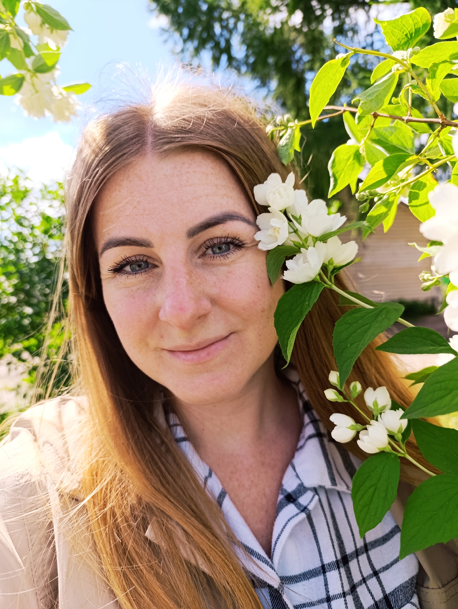 An outdoor, close-up, eye-level, medium-shot photograph captures a fair-skinned woman with long, reddish-blonde hair, facing the camera and smiling. She has light blue eyes, a smattering of freckles across her nose and cheeks, and is wearing a black and white plaid shirt and a light-colored jacket. A branch of white jasmine flowers with green leaves is positioned in the foreground, framing the right side of her face and partially obscuring her hair. The background is blurred, featuring green foliage and a bright blue sky with some clouds. A wooden structure is faintly visible in the upper right portion of the background. The lighting suggests a sunny day.