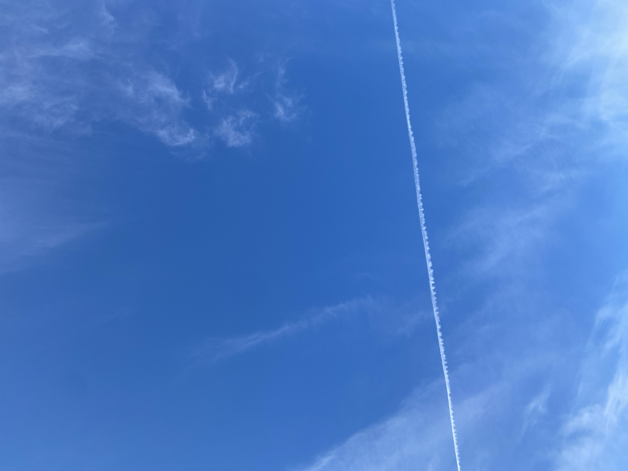 A photograph captures a clear blue sky with wispy white clouds. A prominent contrail, formed by an aircraft, stretches diagonally across the frame from the bottom left to the upper right. The contrail appears as a thin, broken line of white, suggesting it is dissipating. The overall environment is that of an open sky, typical of rural or semi-rural areas like Байқоныс ауылдық округі, Kazakhstan, where clear skies and aircraft activity are common. No people or specific structures are visible, emphasizing the vastness of the sky as the primary subject.