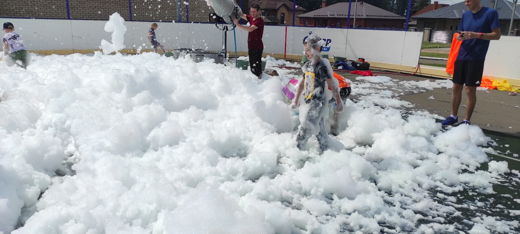 This is a full shot, eye-level outdoor photograph of a foam party taking place in the city of Noviy, Russia. In the foreground, a dense layer of white foam covers the ground, with several children partially submerged in it. A young boy in a dark t-shirt and grey pants is the most prominent child, with a colorful inflatable toy near his left arm. To his right and behind him, another child in an orange shirt is visible. In the middle ground, a man in a red shirt and black pants operates a foam machine, directing a stream of foam. Another child in a patterned shirt is walking away from the foam machine towards the left side of the frame. In the background, a white barrier with blue trim lines the area, and beyond that, buildings with tiled roofs and trees are visible. On the right side of the image, a man in a blue t-shirt, black shorts, and blue athletic shoes stands near the foam, holding a mobile phone. An orange safety vest is visible on the ground behind him, along with a yellow object. The ground surface appears to be a dark, possibly asphalt or rubberized court. The lighting suggests it is daytime with bright sunlight.