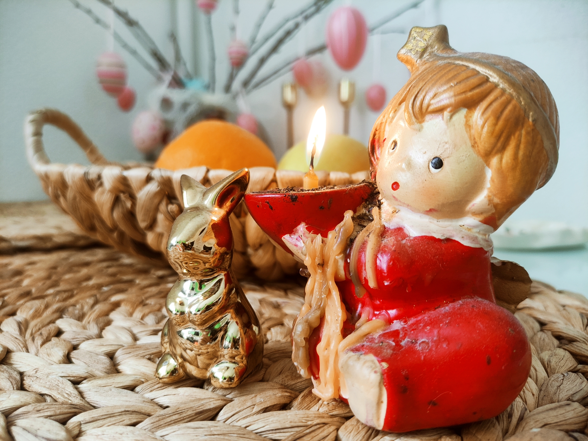 The image features an Easter-themed still life. A woven mat of light brown material serves as the base for the arrangement. A wicker basket filled with an orange and a yellow fruit sits to the left. The background shows decorative tree branches adorned with pastel-colored Easter eggs.
Foreground elements include a gold-colored rabbit figurine and a red and beige figurine of a girl holding a candle. The candle is lit, with its flame visible. The scene is photographed indoors under natural light. The location is Unknown, Unknown.