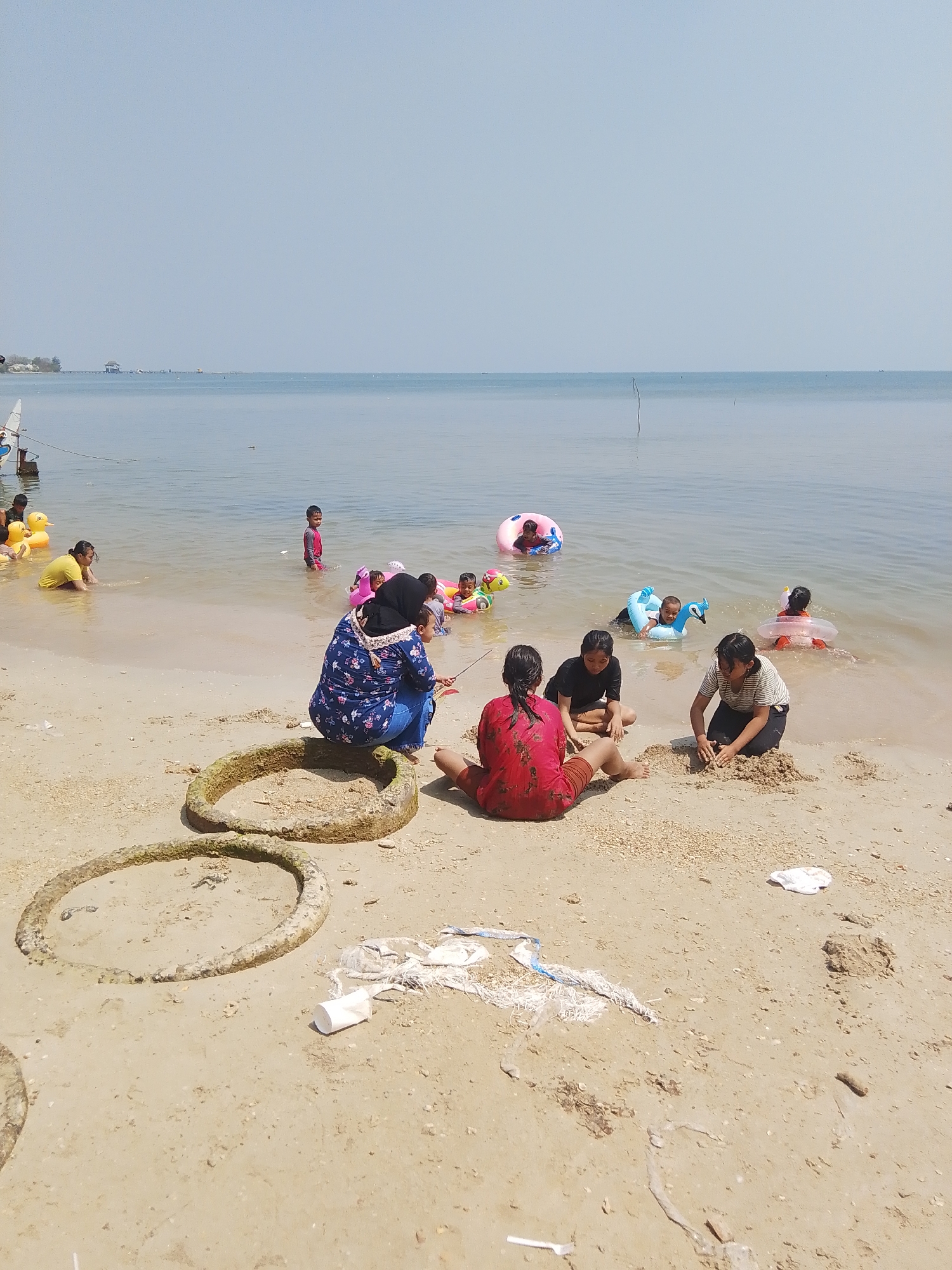 In the city of Pejagan, Indonesia, a group of people are enjoying a day at the beach. The scene depicts a sandy shoreline meeting calm, shallow water under a clear blue sky. Several children are playing in the water, some with inflatable rings, while others, including a few adults, are on the sand. Two large, circular concrete objects, possibly remnants of old construction or infrastructure, are placed on the sand, and one appears to have sand piled inside it, suggesting it's being used as a play area. A woman in a dark blue and white floral top and blue jeans is sitting on the sand, holding a child, facing away from the camera. Two other women, dressed in black t-shirts, are sitting on the sand and building a sandcastle. In the background, the serene ocean stretches to the horizon, with a faint silhouette of a building and possibly a pier on the left side of the image. The overall atmosphere is one of leisure and recreation.