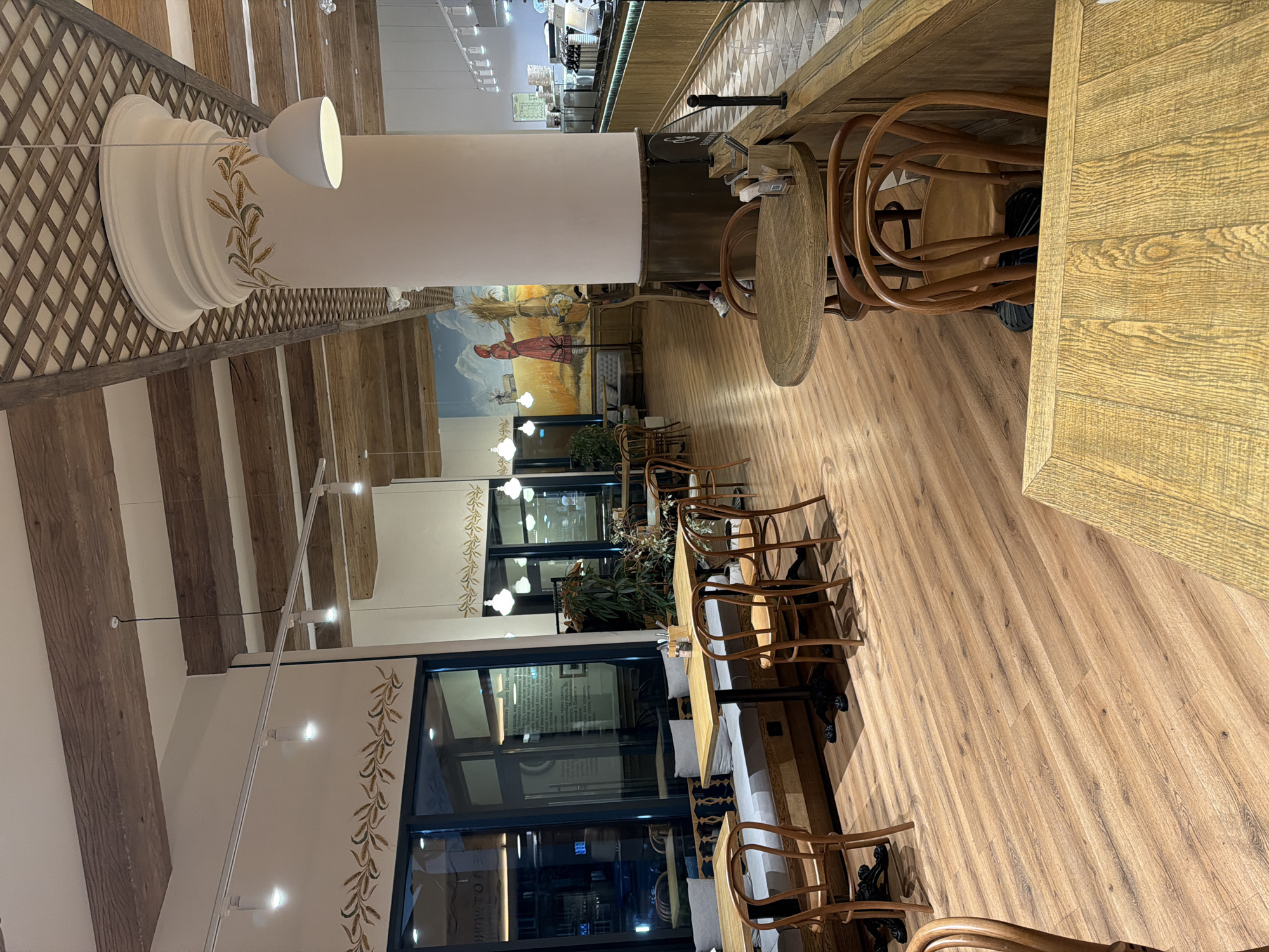 The image shows the interior of a restaurant or cafe. The location is identifiable as Izhevsk, Russia, based on the Cyrillic lettering visible on exterior signage through the windows. The dining area features wooden floors and tables with chairs. Some tables have cushioned bench seating along the wall. A large cylindrical light fixture hangs from the ceiling, decorated with wheat-like designs. A mural adorns the wall in the background, depicting a pastoral scene. The space appears to be empty of patrons at the time the photo was taken. Windows line one side of the room.