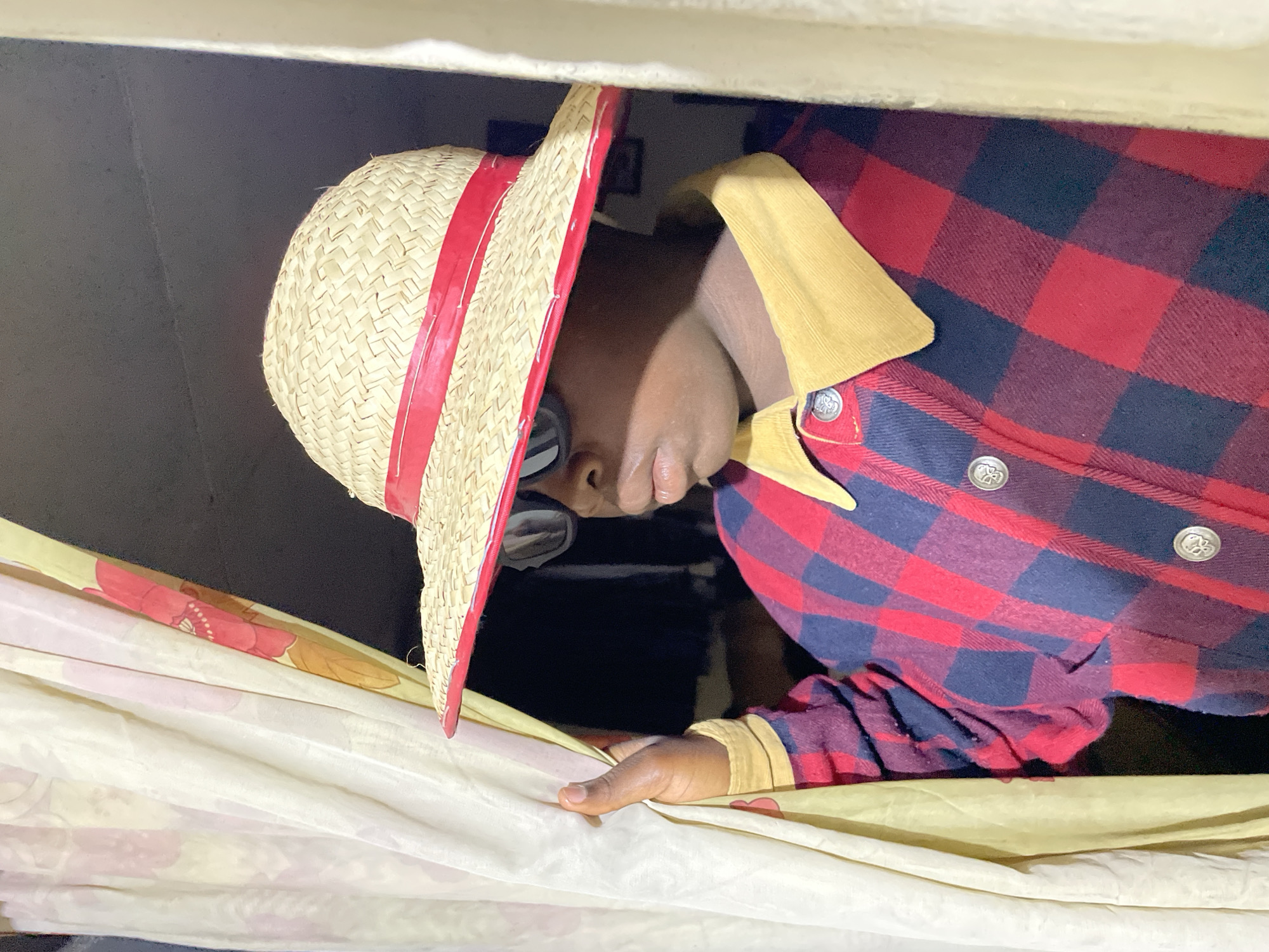 A person wearing a straw hat with a red ribbon, glasses, and a red and blue plaid shirt with a tan corduroy collar is partially visible. The person is positioned close to a white floral patterned curtain. The environment appears dimly lit, and based on the context of Jos, Nigeria, the scene could be indoors. No specific city details are discernible.