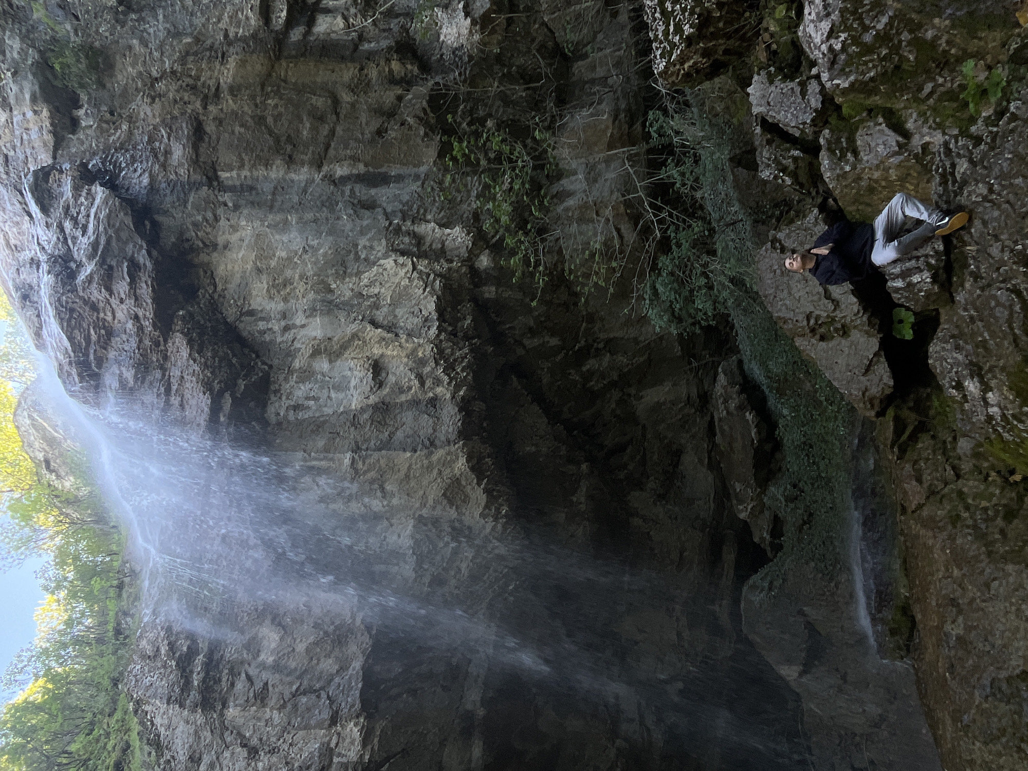 The image depicts a waterfall cascading down a steep, rocky cliff face. Lush green foliage clings to the rocks on either side of the falling water. On the right side of the image, a person is perched on a ledge, appearing to be observing the waterfall. The location is likely near Veselinovo, Bulgaria, given the prompt. The environment is characterized by rough, natural textures of the rocks and the soft, blurred movement of the water.