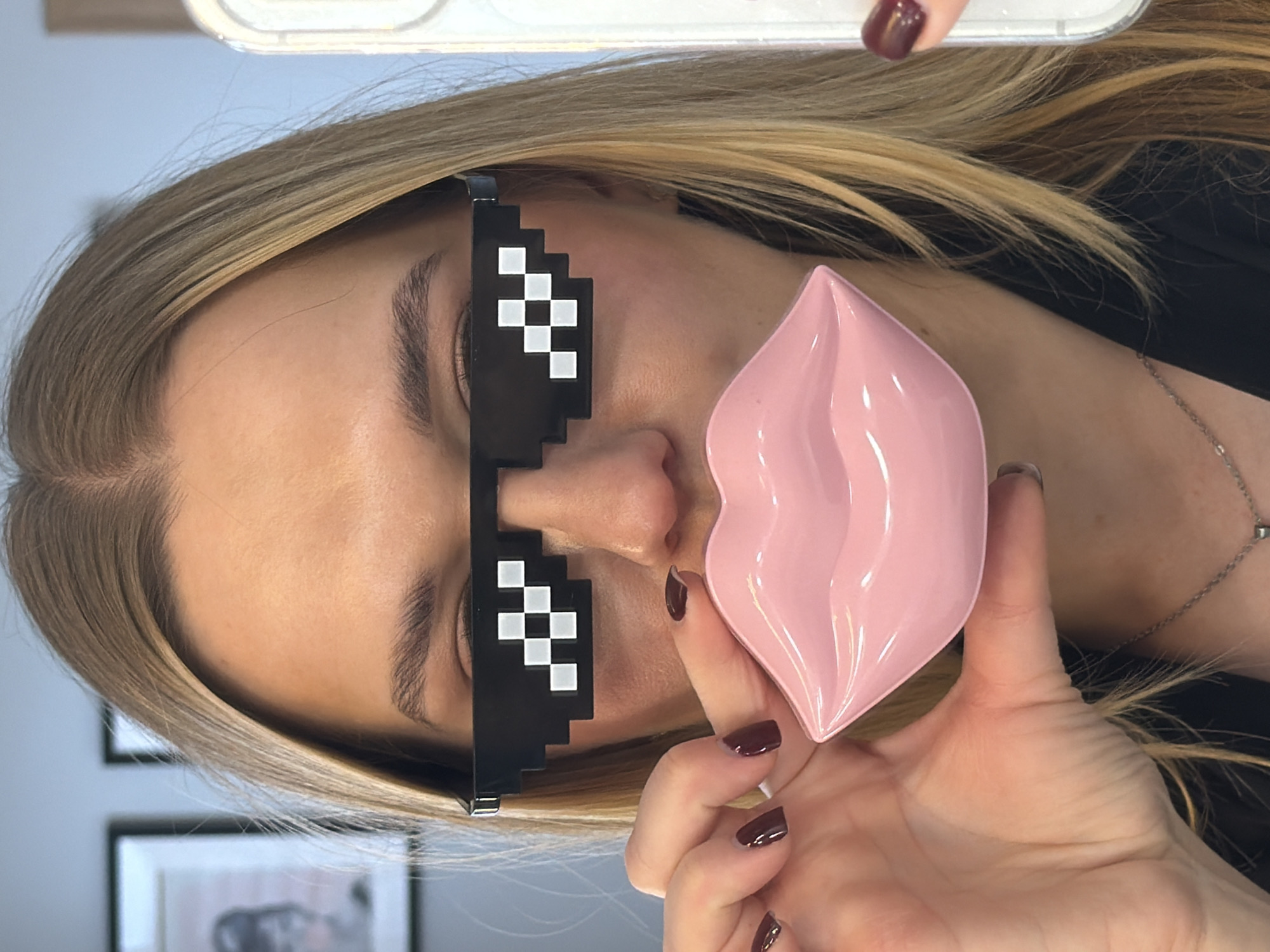 A woman with blonde hair and pixelated sunglasses is holding a pink, lip-shaped object. She is wearing dark nail polish and a silver necklace. The background includes framed artwork and a white surface, suggesting an indoor location. While specific details do not confirm the city of Riga, Latvia, the image depicts an interior scene that could be anywhere.
