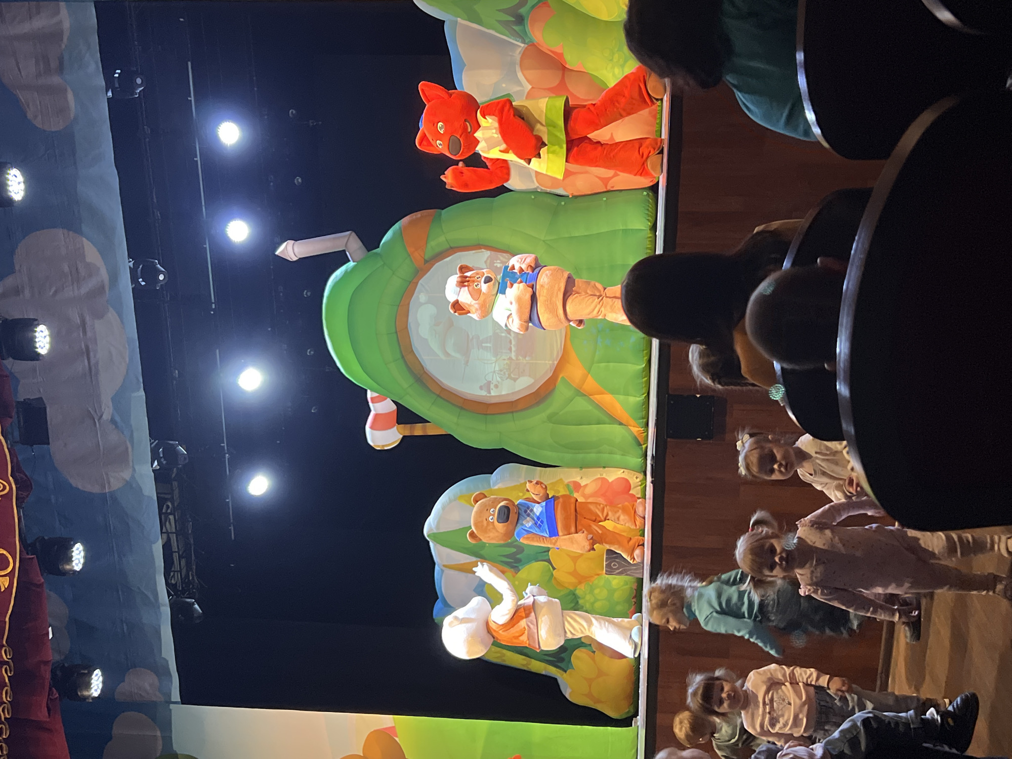 The image captures a performance stage with inflatable cartoon characters. The characters, including a red fox, a dog, a bear, and a white rabbit, are positioned against a backdrop of green, yellow, and blue inflatable elements that resemble foliage and other natural features. Several spotlights illuminate the scene from above.
In the foreground, a number of young children are seated in an auditorium setting. Some of them are looking up at the stage. Behind them are rows of theatre-style seats.
The location is likely Chaikovsky, Russia, based on visual clues and general knowledge. The image depicts a children's theater performance.