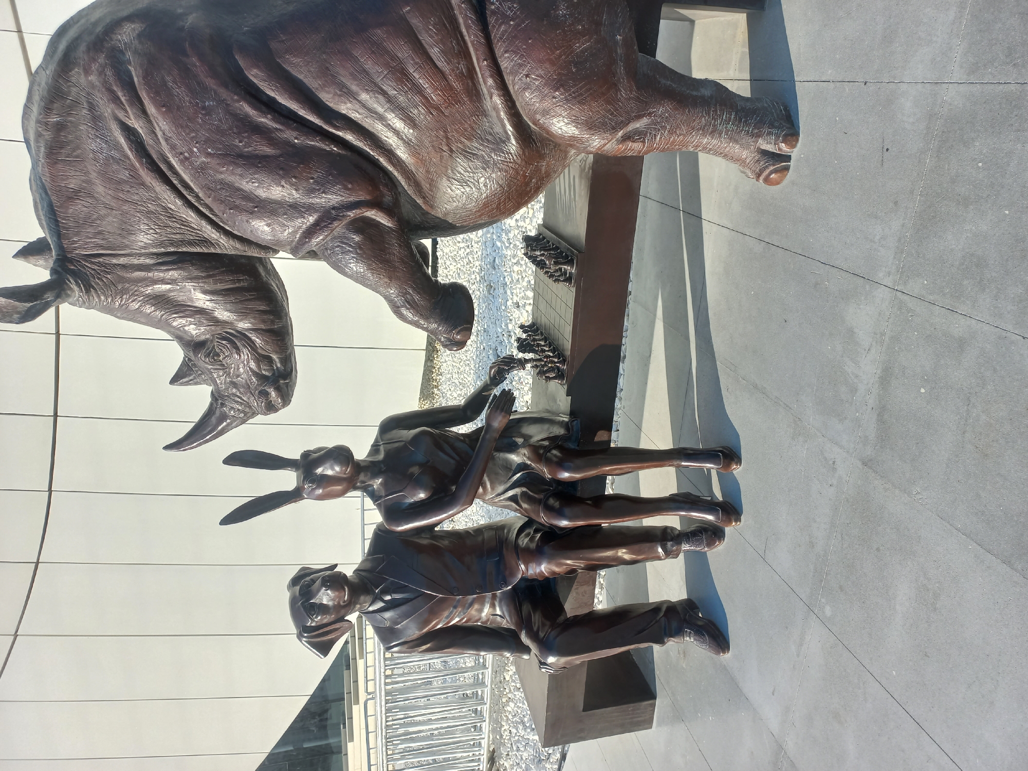 This is a bronze sculpture depicting a dog-headed man in a suit, a rabbit-headed woman in a dress, and a rhinoceros. They are seated on a bench in front of a chess board. The dog and rabbit figures are playing chess. The rhinoceros is positioned to the right of the bench, looking towards the chess board. The sculpture is located in Baku, Azerbaijan, possibly in a public park or plaza, with a modern building in the background. The ground is paved with large square tiles, and there is a low white fence behind the bench. The scene is illuminated by sunlight, casting shadows on the ground.