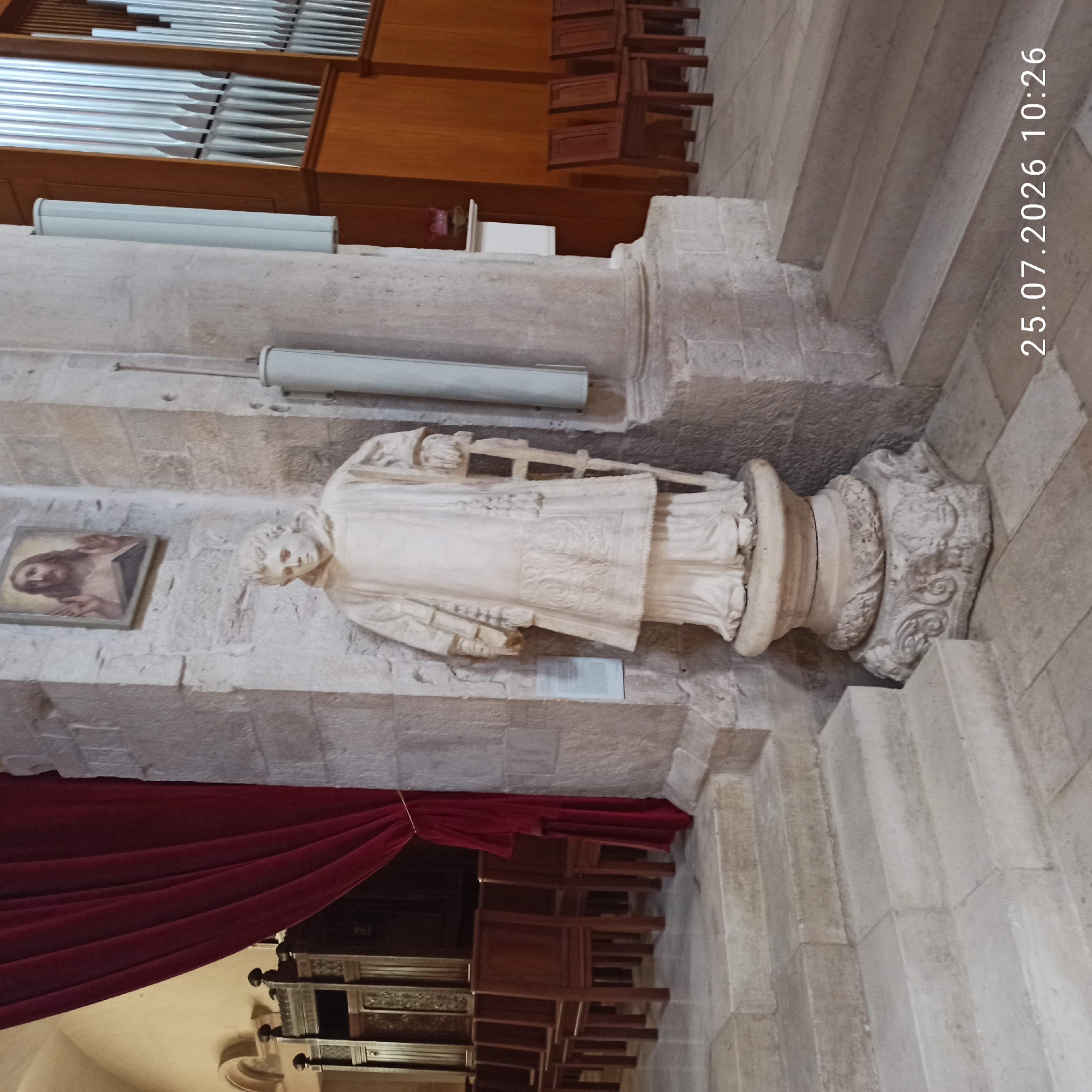 The image shows the interior of a church in Ruvo di Puglia, Italy, likely the Basilica di Santa Maria Assunta. In the center of the frame is a stone statue of a robed figure, possibly a saint or cleric, standing on a decorative pedestal. The statue is white and appears to be carved from marble or a similar stone. To the left of the statue, a red velvet curtain hangs beside a dark wooden ornate cabinet or choir stall. Behind the statue, a framed religious painting of Jesus is mounted on the stone wall. To the right of the statue, a large pipe organ with silver pipes is visible against a wooden background. The environment is constructed from stone blocks and has a light-colored tiled floor. In the foreground, stone steps lead up towards the main area of the church. The bottom right corner of the image contains a timestamp: "25.07.2026 10:26".