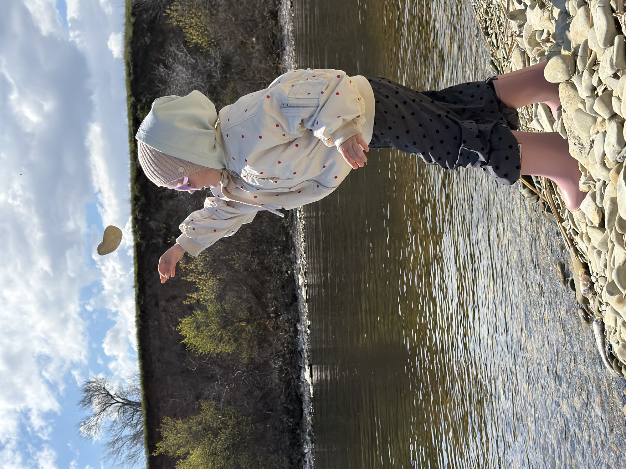 A young child stands on a pebble beach beside a body of water. The child wears pink boots, a dark skirt with a light-colored jacket with red polka dots. A light-colored hooded sweatshirt and a knit hat are worn on the child's head. The child is in the action of throwing a stone. On the other side of the water is a vegetated embankment, with the sky visible in the background. This is in Kamyshin, Russia.