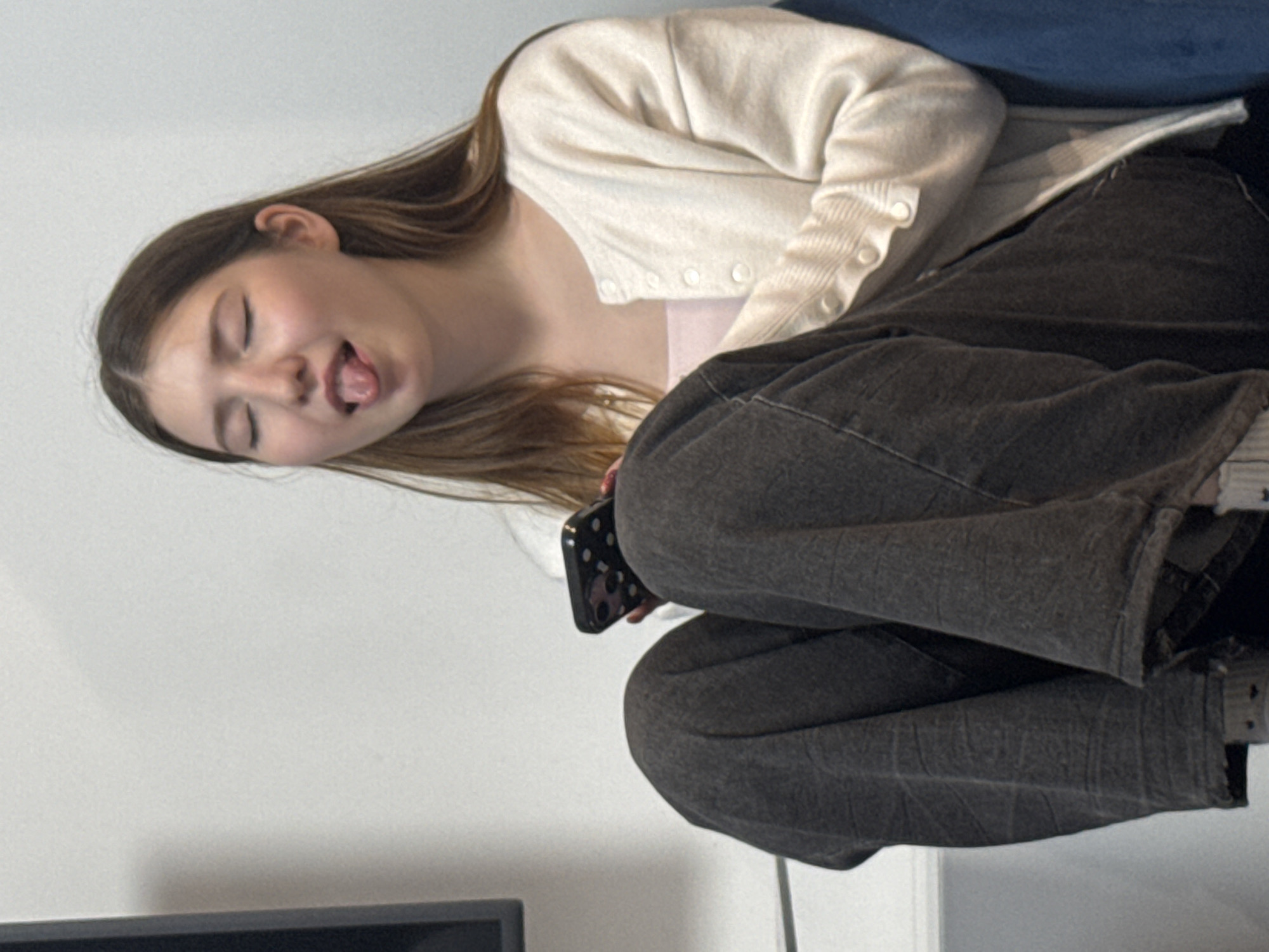 A young person with long, brown hair is sitting in a chair with their legs crossed, holding a smartphone. They have their eyes closed and their tongue sticking out. They are wearing a light-colored cardigan over a pink top and dark-colored pants. The background is a plain white wall.