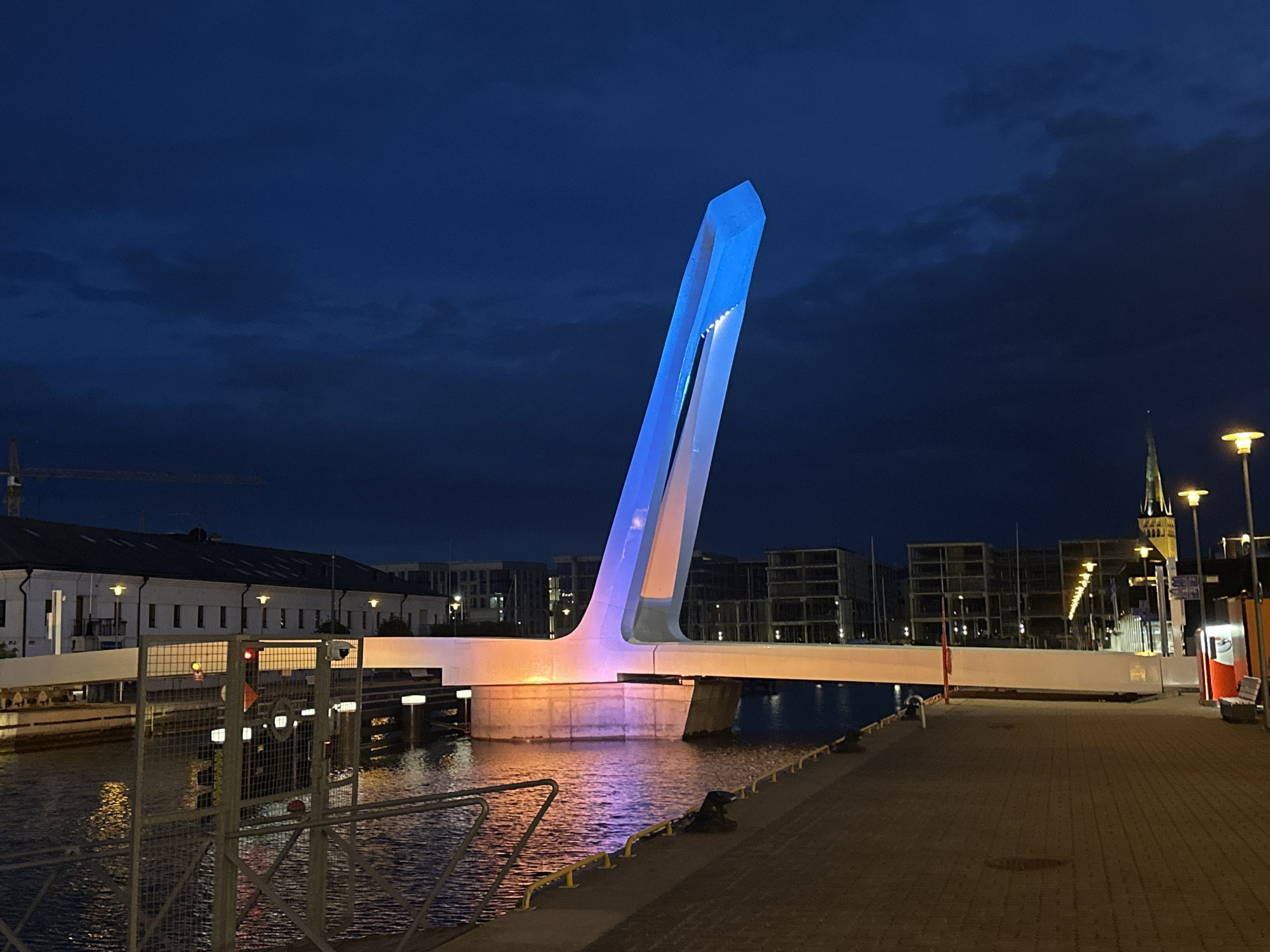 The image depicts a modern bridge illuminated with blue and orange lights at dusk in Tallinn, Estonia. The bridge is a prominent architectural feature, with a tall, angular structure that appears to be a pylon or support. The water below reflects the vibrant lights of the bridge and the surrounding urban landscape. In the background, there are buildings, streetlights, and a tower with a spire, suggesting a harbor or waterfront area within the city. The sky is a deep blue, indicating the transition from day to night, with some subtle cloud formations. The scene conveys a sense of urban activity and modern design within a historical city.