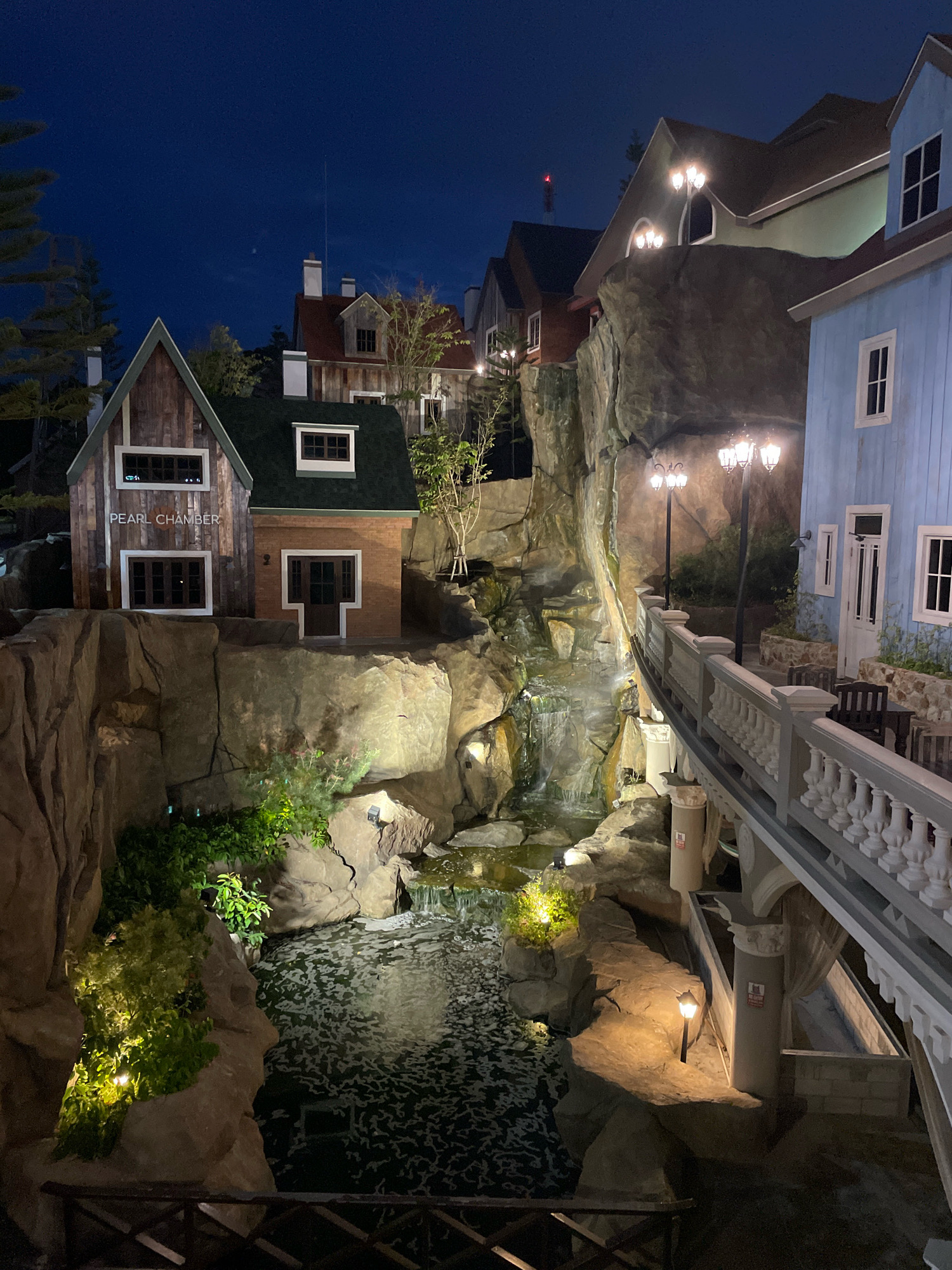 This is a nighttime photograph of an artificial landscape scene in Pattaya City, Thailand, designed to resemble a European village with a waterfall and stream. The scene features several small, elaborately decorated buildings with a rustic, wooden facade on the left, labeled "PEARL CHAMBER," and a light blue building on the right. A walkway with a decorative balustrade spans across the scene, overlooking a man-made stream with a small waterfall cascading down rocks. The area is illuminated by warm-toned artificial lights, including lampposts and spotlights highlighting the greenery and rock formations. There are no people visible in the frame. The overall impression is one of a meticulously crafted, picturesque tourist attraction.
