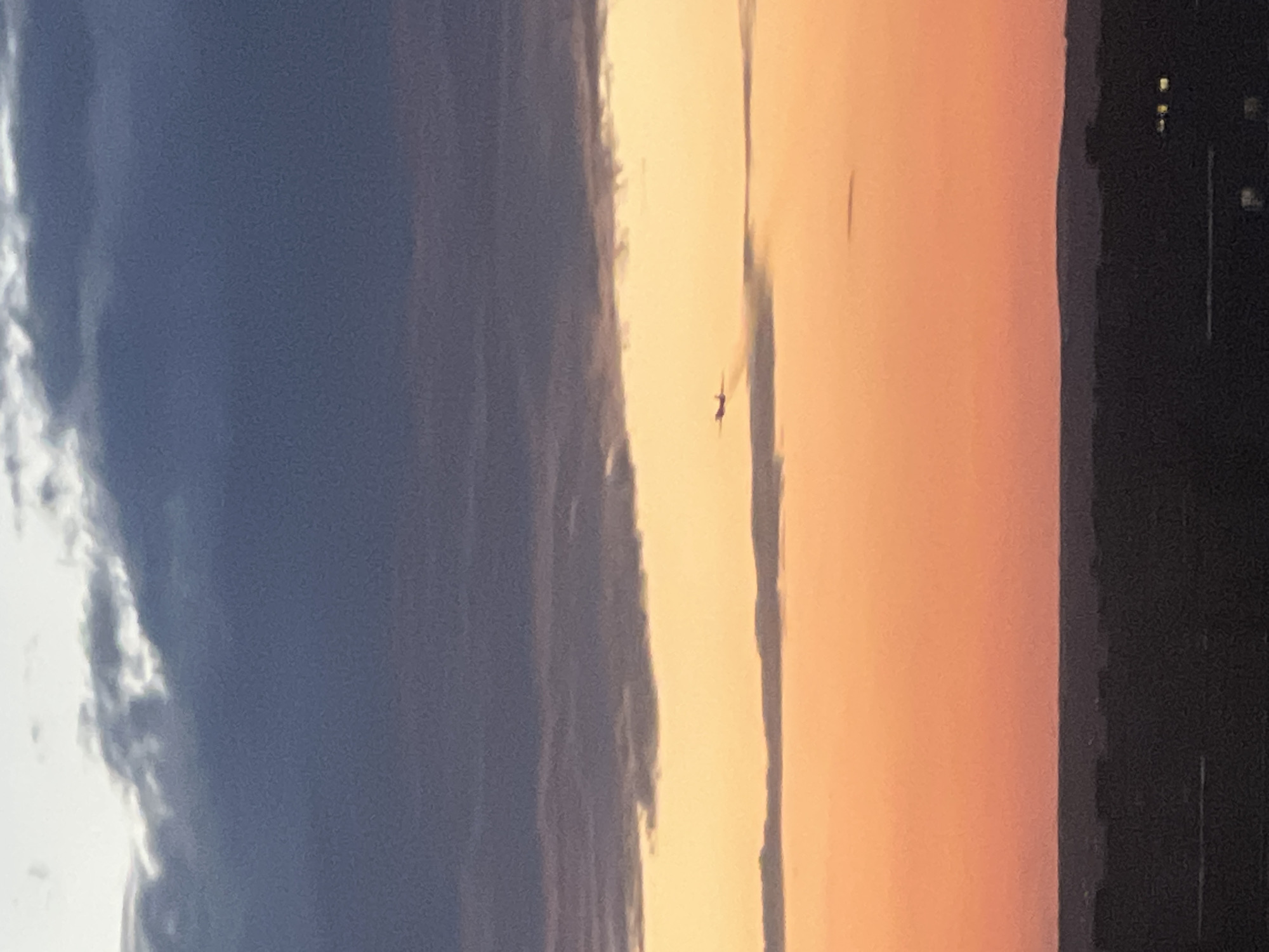 An airplane is seen flying in the sky above a city at dusk. The sky is filled with dramatic clouds in shades of dark blue, gray, and orange, indicating the setting sun. The airplane is a small silhouette against the bright orange and yellow hues of the lower sky, leaving a faint contrail. Below the sky, a dark silhouette of the city skyline is visible, with some distant lights suggesting urban activity. The overall scene captures a moment of transition from day to night, with an aircraft adding a dynamic element to the atmospheric landscape.
