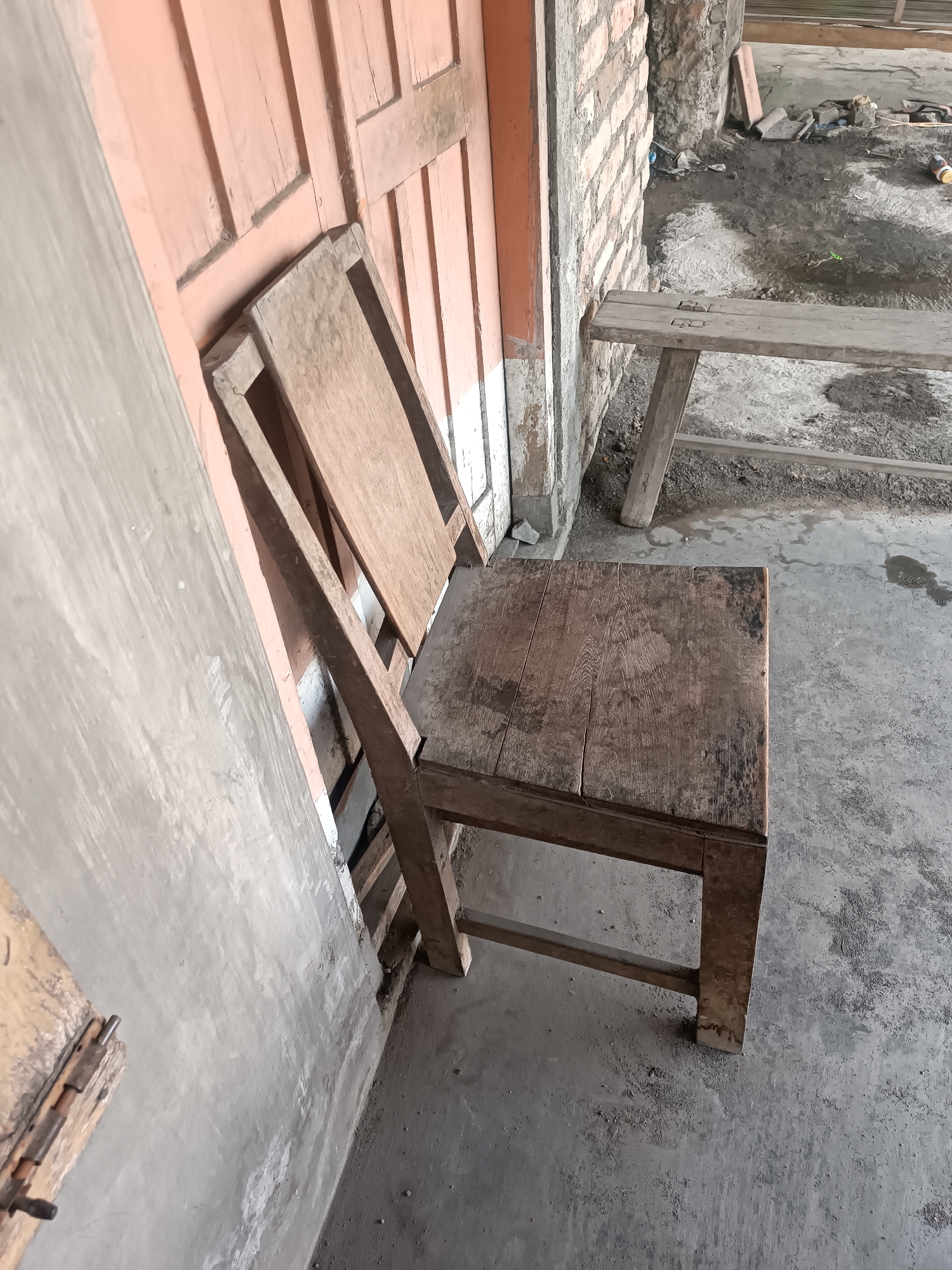 This is a medium-shot, eye-level, outdoor photograph of a wooden chair placed next to a building wall. The building has a light pink wooden door and a rough brick wall. To the right of the chair is a simple wooden bench. The ground is concrete and appears wet in places. Debris, including plastic bottles and other refuse, is scattered in the background. The scene appears to be an unkempt area, possibly a construction site or an outdoor storage space. The lighting is diffuse, suggesting an overcast day or shade. The image is likely taken in the city of Jetis, Indonesia, based on the context provided, implying a local setting.