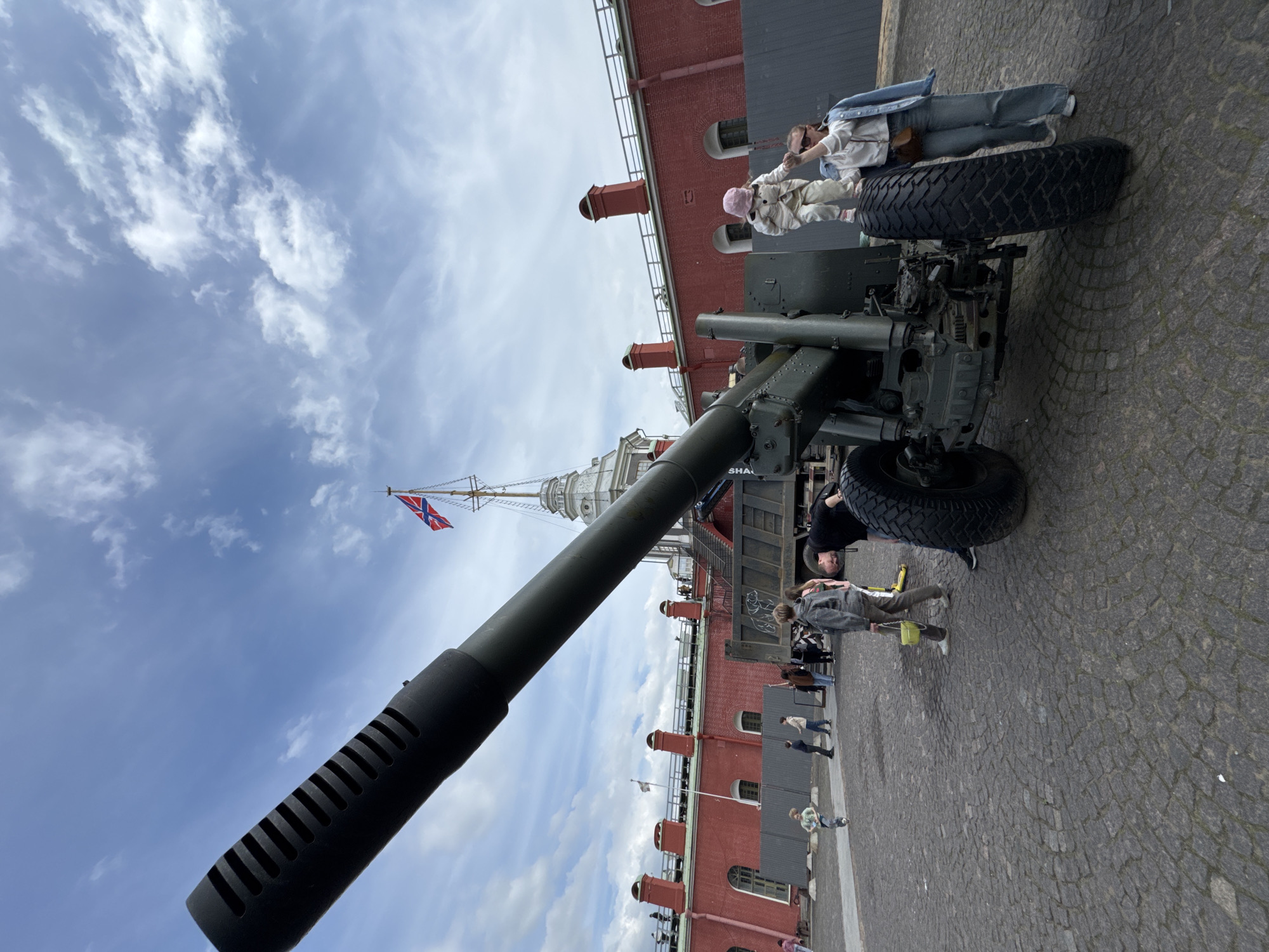 This image depicts an outdoor scene at a historical site, likely in Saint Petersburg, Russia, under a partly cloudy sky. The central subject is a large, dark green artillery gun, possibly a howitzer or field gun, resting on sizable rubber-tired wheels. Its long barrel, equipped with a muzzle brake, extends prominently into the foreground.
Several people are present. To the right of the cannon, an adult wearing a light denim jacket and jeans is seen with a child in light-colored clothing and a pink hat, appearing to assist the child near the gun's large wheel. Another individual in a black top is bending down near the back of the artillery piece. Other people are casually walking on the cobblestone ground in the background.
Behind the artillery, a red brick building with arched windows and distinctive red decorative elements along its roofline is visible. Above this building, a tall, white spired tower, characteristic of Saint Petersburg architecture, rises into the sky. A flag, appearing to be a Russian naval ensign (blue St. Andrew's cross on a white background), flies from a mast adjacent to the spire. The ground is paved with cobblestones, and the sky is a light blue with scattered white clouds, suggesting pleasant daytime weather. Part of a dark green truck is visible behind the gun, with the word "SHAG" partially readable on its side. The scene conveys a relaxed atmosphere of tourists visiting a historical military exhibition or fortress.