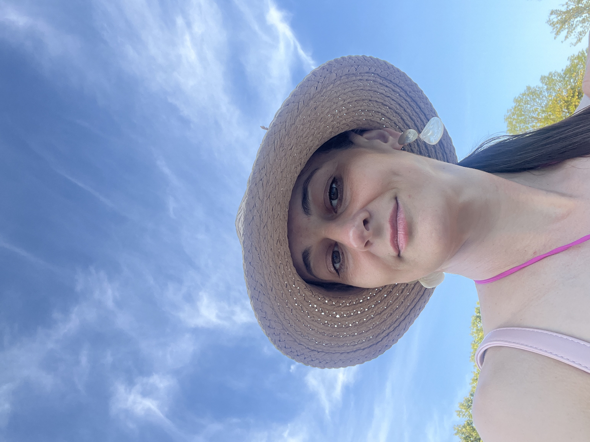 A close-up, low-angle, selfie-style shot captures a woman wearing a straw hat and a pink bikini top against a backdrop of a bright blue sky with wispy white clouds. Her face is centered in the frame, with her eyes looking slightly off-camera. She has on distinctive earrings, one of which is partially visible. To the right of her face, her dark hair is pulled back, and to the left, a portion of her left shoulder and the strap of her bikini are visible. In the background, the tops of green trees can be seen against the sky, suggesting an outdoor setting, possibly during daytime.