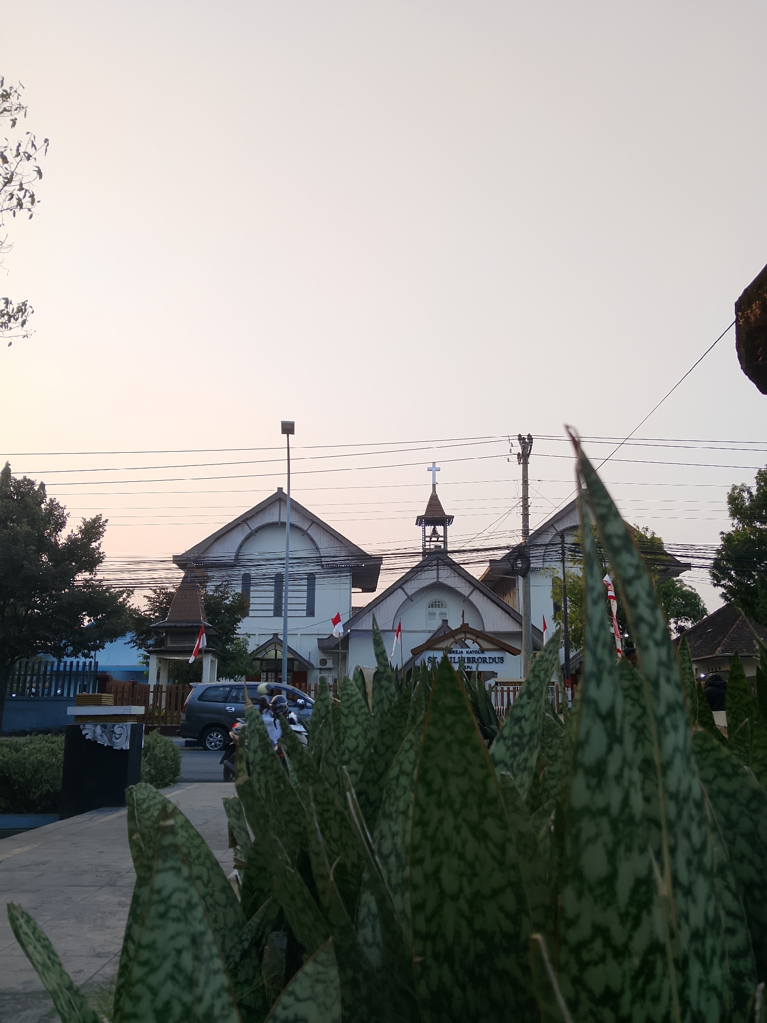 The image depicts a building in Balun, Indonesia, identified by signage as "GEREJA KATOLIK ST. ILISBORDUS" (Catholic Church of St. Ilisbordus). The building has a white facade with dark gray trim and a pitched roof. A cross is visible on top of a steeple, and a bell tower is also present. In front of the church, there is a paved area with a sidewalk, a fence, and landscaping featuring snake plants in the foreground. A gray minivan and a motorcycle with a rider are on the road in front of the church. Indonesian flags are displayed on poles near the church entrance. The sky is a light, hazy orange, suggesting it may be dawn or dusk. Utility poles and power lines are visible against the sky, running over the church and surrounding area.