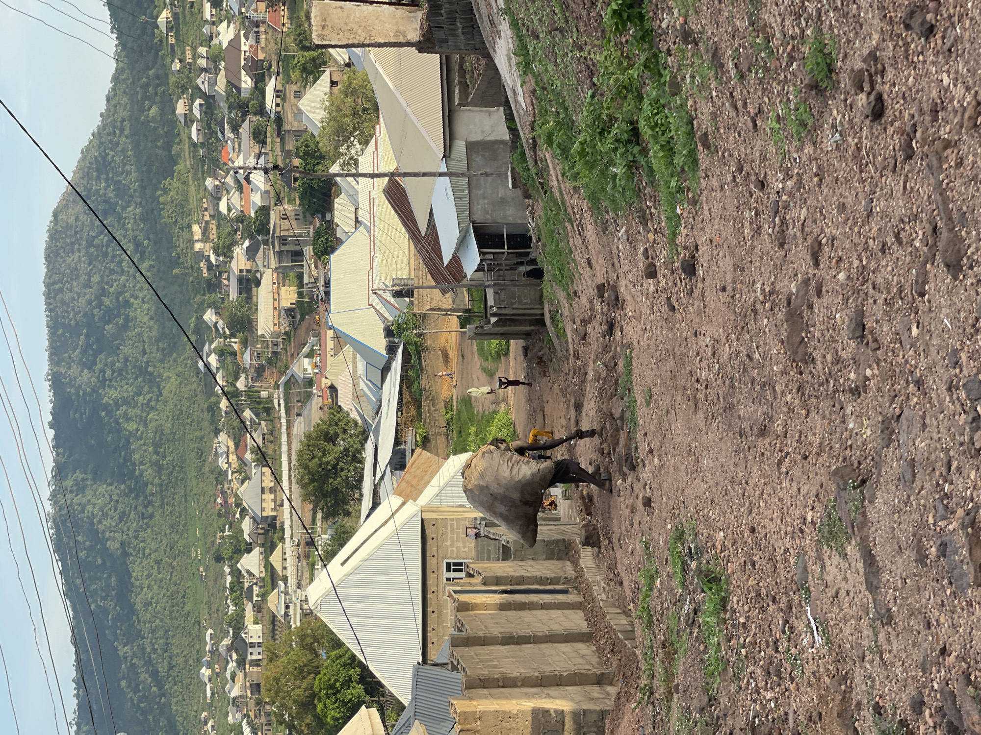 The image captures a rural or semi-urban scene in Lokoja, Nigeria, set on a hillside under bright daylight. A dirt path, made of reddish-brown gravel and earth with sparse green vegetation on its edges, slopes downwards from the foreground towards a settlement.
In the mid-ground, a person walks down this path, carrying a large, heavy-looking sack on their back, held by straps over their shoulders. Further down the path, two smaller figures are visible; one appears to be pushing a wheelbarrow or similar cart.
The setting is characterized by numerous houses built into the sloping terrain. These structures primarily feature unpainted cinder block walls and corrugated metal roofs, mostly silver or light-colored, though some have a rusty, brownish hue. Many of the buildings appear to be under construction or unfinished. Overhead, power lines stretch across the scene.
In the background and to the left, a large, lush green mountain covered in dense vegetation dominates the horizon, contrasting with the built environment. The sky is clear and bright, indicating a sunny day. There is no visible text in the image.