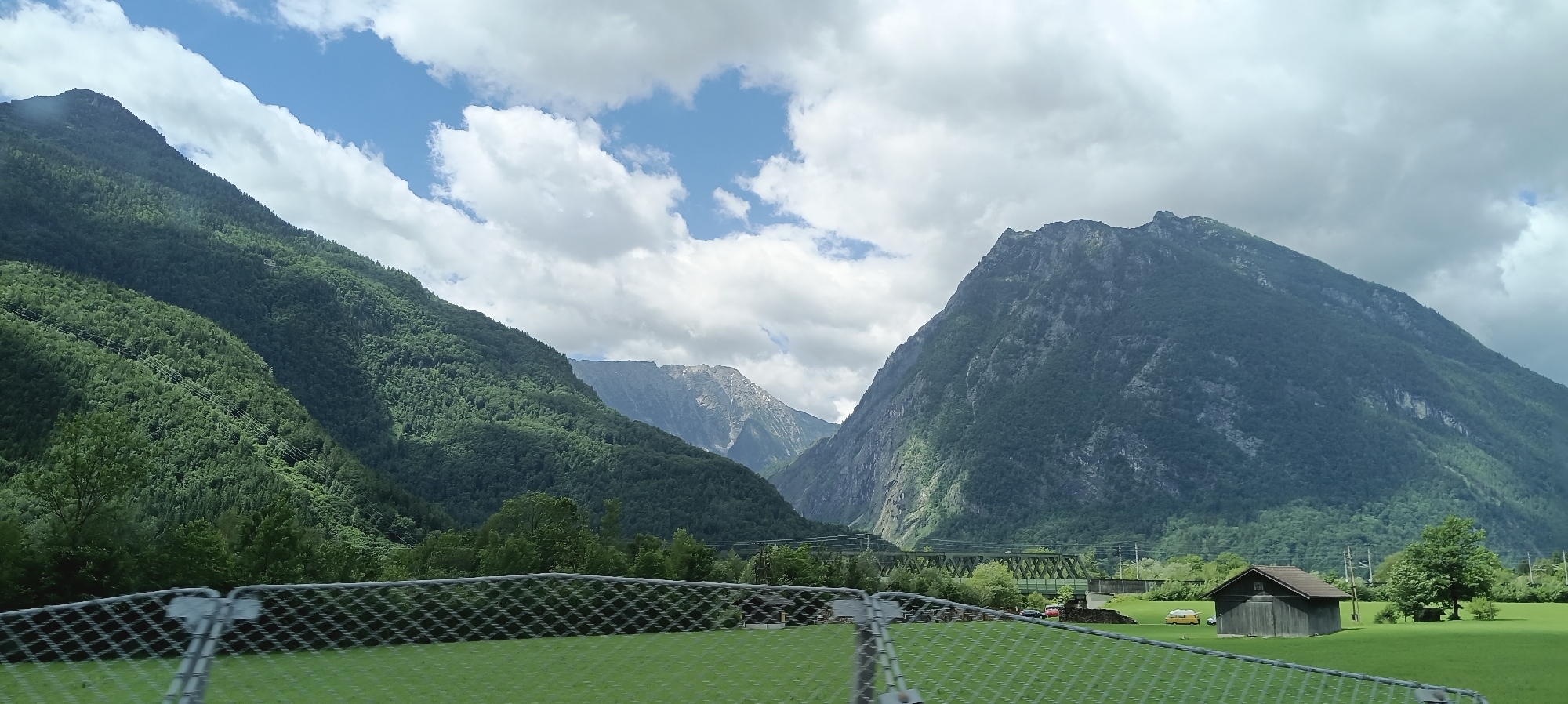 The image depicts a scenic mountain landscape in Torren, Austria. In the foreground, a metal chain-link fence is visible, partially obscuring a lush green grassy field. Beyond the field, a small, dark wooden shed with a sloped roof stands near a mature tree. In the middle ground, a green metal railway bridge spans across a valley, with a train track visible. The background is dominated by imposing, verdant mountains that rise steeply towards a sky filled with white, fluffy clouds. The slopes of the mountains are covered in dense green forest. In the distance, other mountain peaks are visible, some with rocky, exposed faces. The overall scene is one of natural beauty and tranquility.