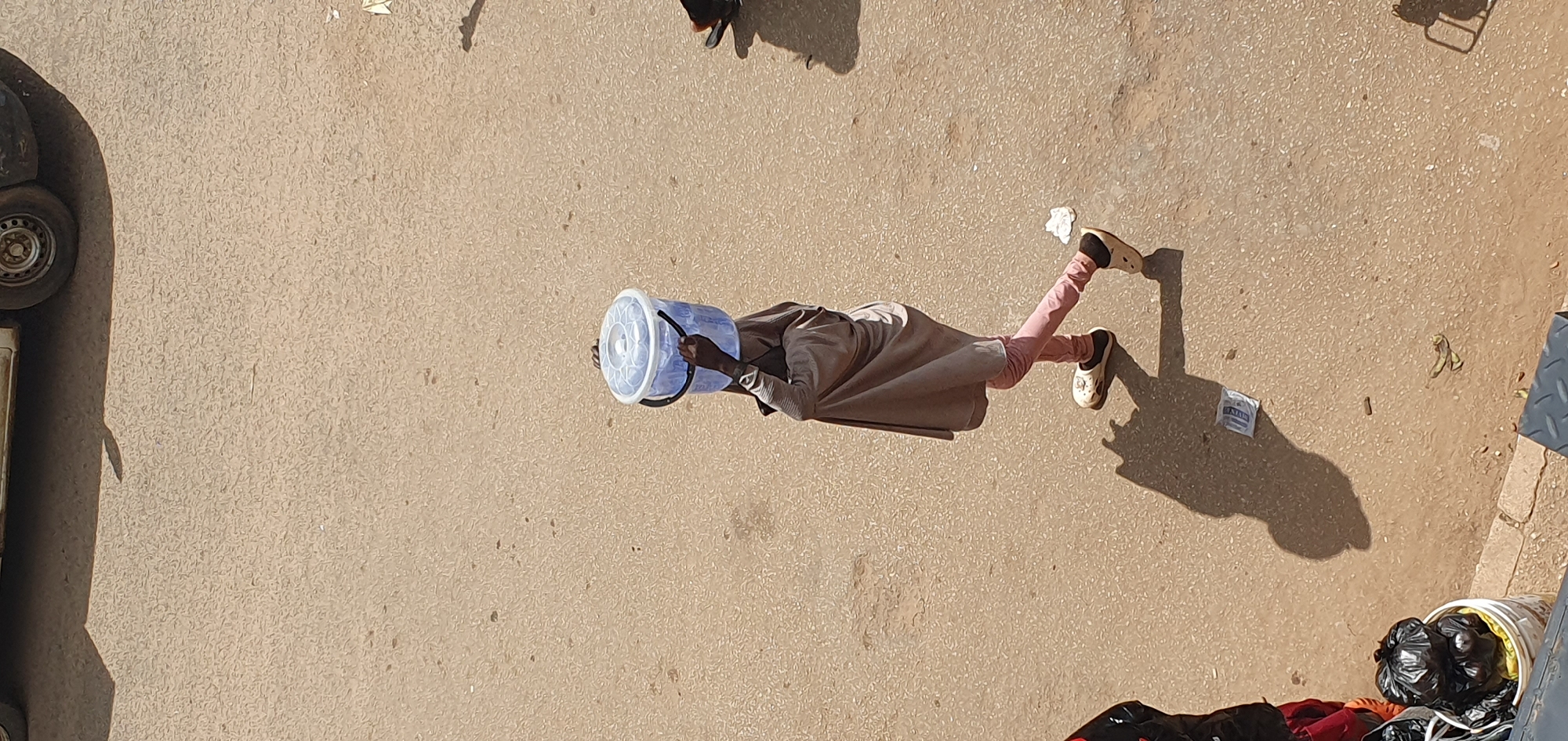 In Bukuru, Nigeria, a person is walking on a paved surface, carrying a large, transparent blue plastic bucket filled with water on their head. The individual is wearing a grey garment that drapes over their shoulders and arms, pink leggings, and light-colored slip-on shoes. Their shadow is cast on the ground to their left. The environment appears to be an outdoor urban setting, with a portion of a car visible at the top of the image, casting its own shadow. A white bucket containing black trash bags is on the left side of the frame. The ground is a textured, light brown asphalt or concrete.
