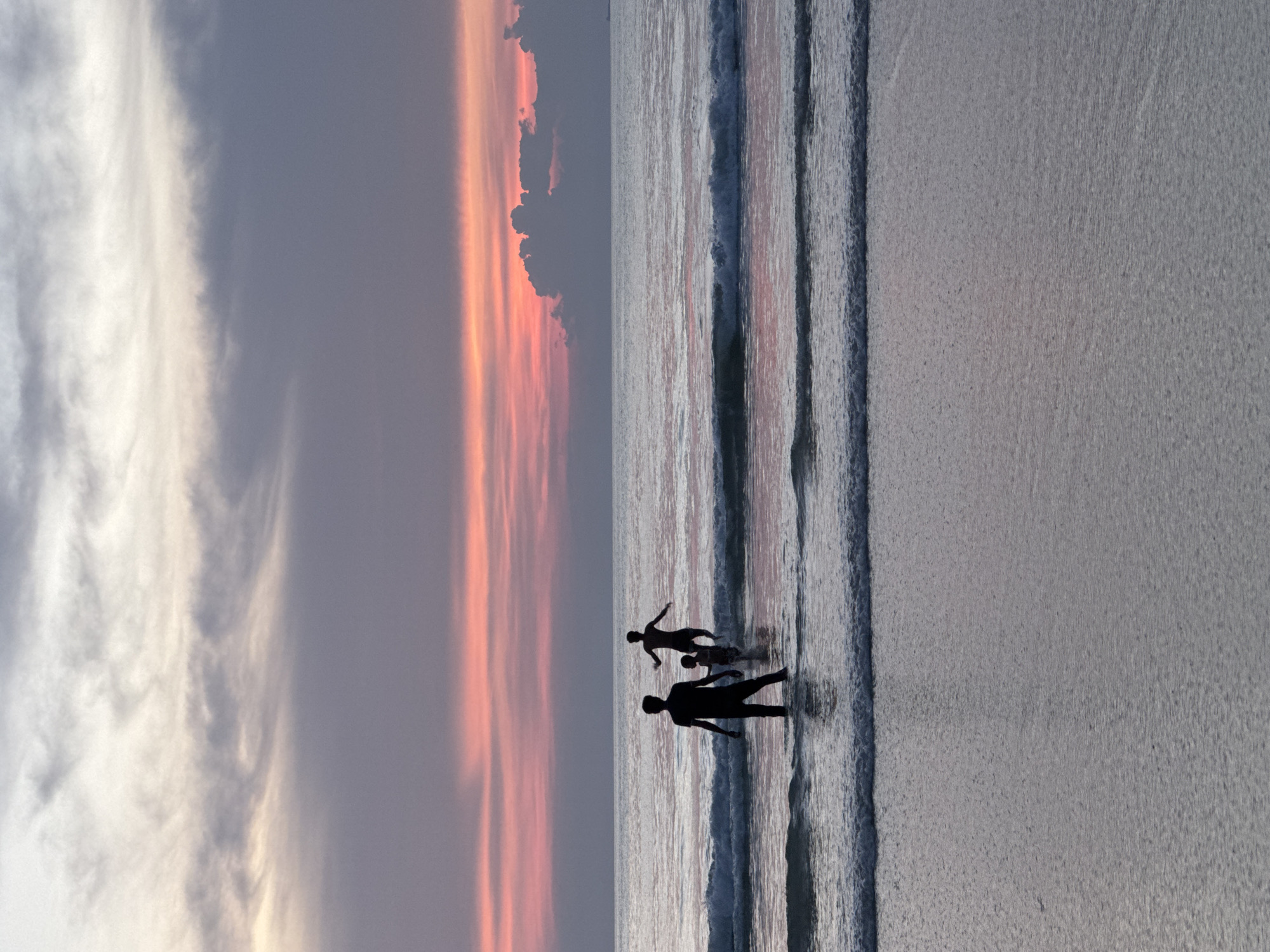 In Patong, Thailand, at dusk, three figures are silhouetted against a tranquil sea and a vibrant sunset. The sky is a canvas of soft purples, pinks, and grays, with the sun having just dipped below the horizon, casting a warm glow. Gentle waves lap at the shore, creating a shimmering effect on the wet sand and shallow water.
In the foreground, a lone figure stands in the water, facing away from the camera, their silhouette dark against the sea. Further out, two other figures are visible. One appears to be an adult with arms outstretched, wading in the water, while a smaller figure, possibly a child, stands close by. The water is calm, with small waves creating ripples around their legs. The scene evokes a sense of peace and familial connection, set against the backdrop of a beautiful tropical sunset.