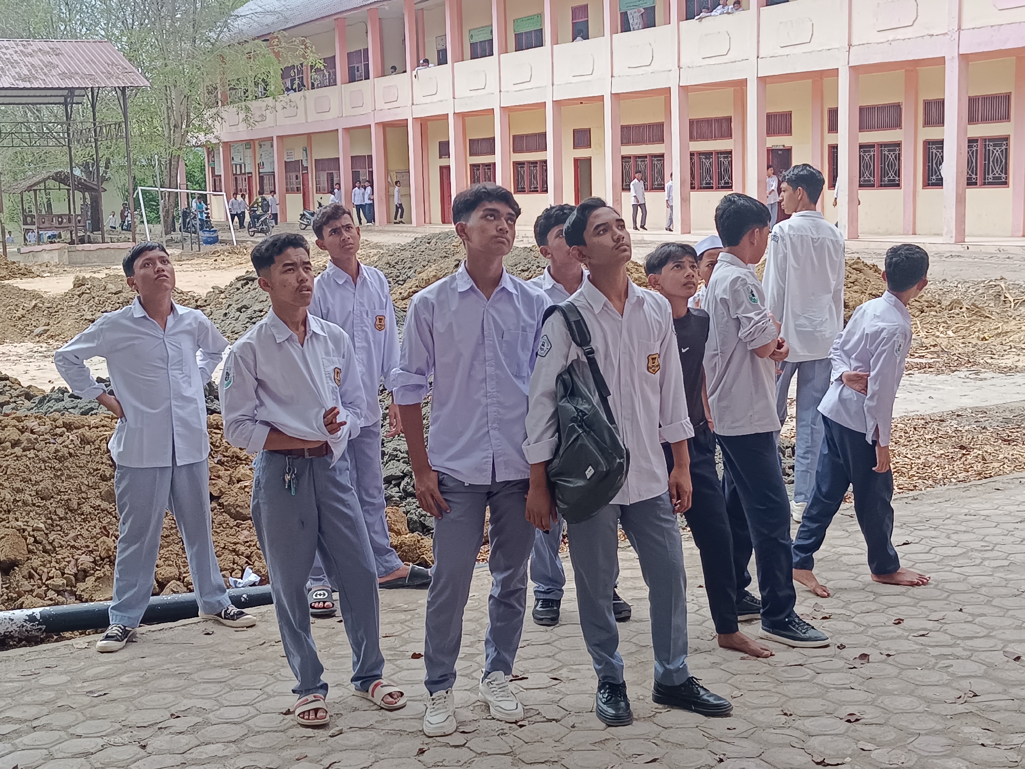 A daytime shot shows a group of approximately ten young men wearing white collared shirts and gray or navy blue pants standing outdoors. The setting appears to be a schoolyard or campus in Lhokseumawe, Indonesia, based on the architecture and clothing style. They are all looking up and in the same direction. Some are wearing shoes or sandals; others are barefoot. A two-story building with multiple windows and columns can be seen in the background. Piles of dirt and construction work are visible in the foreground, possibly indicating ongoing landscaping or renovation. Several other individuals are visible in the background near the building and in another outdoor structure.