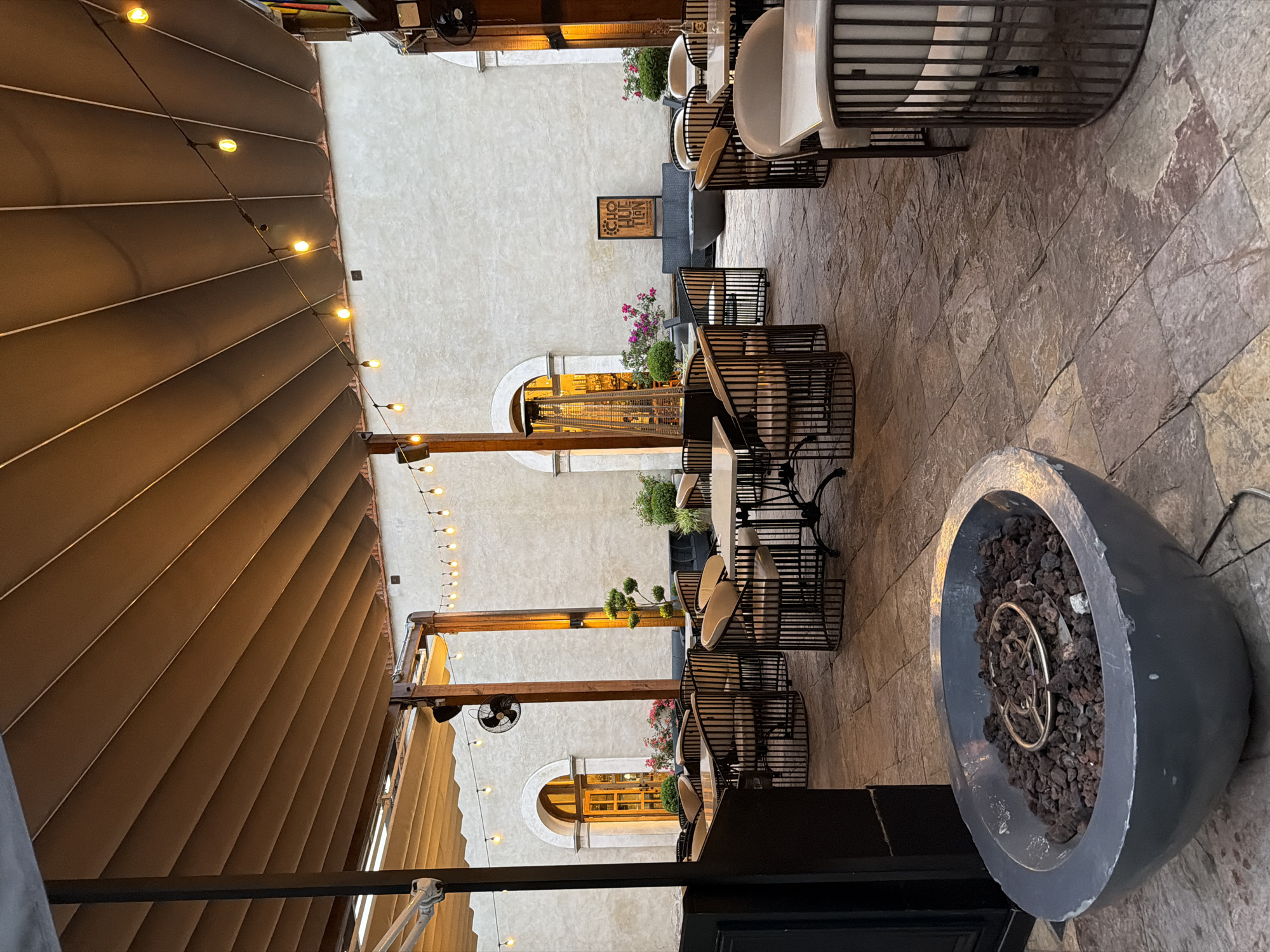 The image shows an outdoor patio setting in Domingo Arenas, Mexico. The space is characterized by stone pavement and a retractable awning overhead, adorned with string lights. Several tables with metal-framed chairs are arranged throughout the area. A large, spherical fire pit sits in the foreground, filled with volcanic rock and a metal ring. The background features a white building with arched windows and various plants.