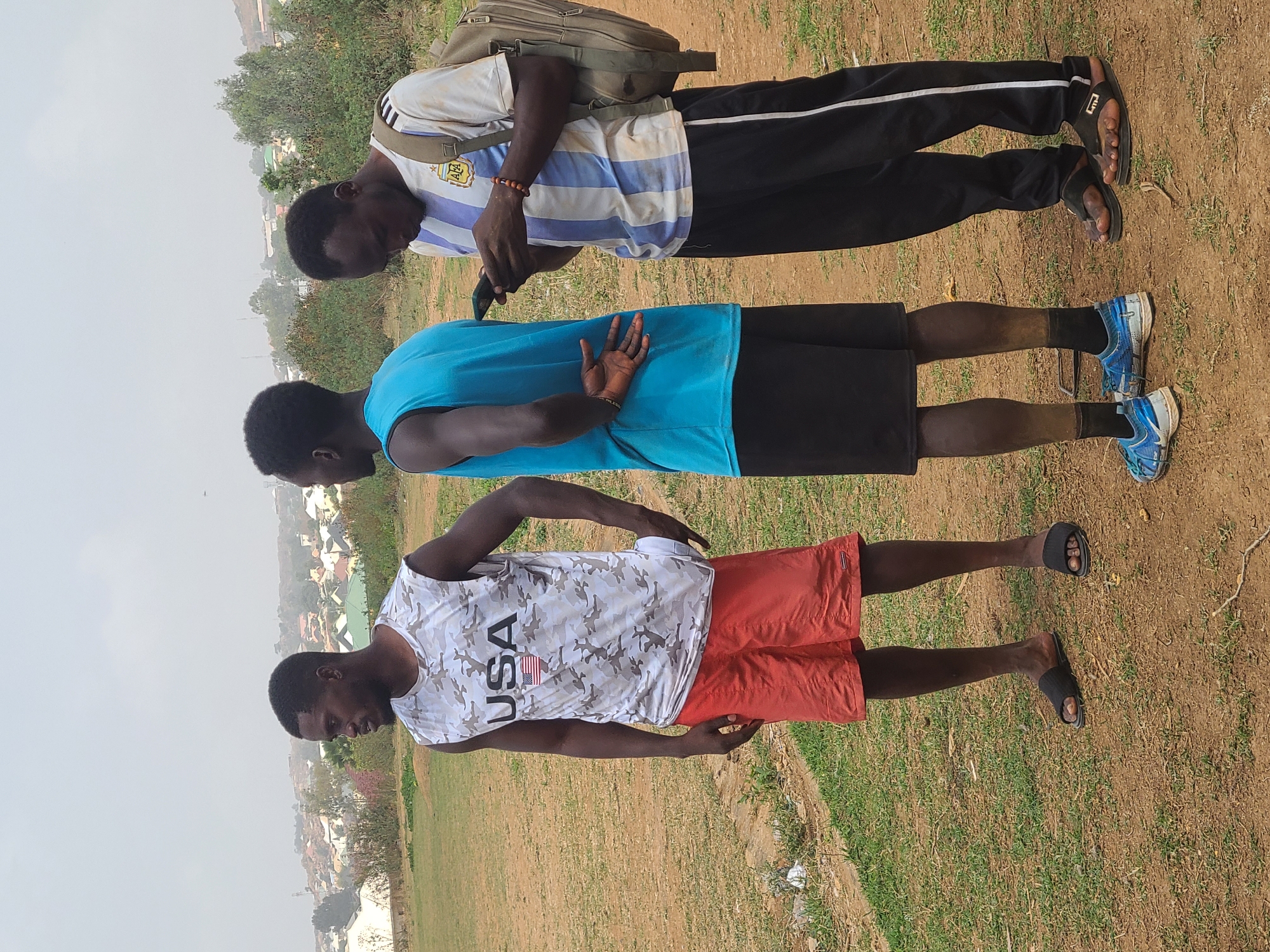 Three African men stand in a field. The man on the left wears a white and blue striped Argentina soccer jersey, black sweatpants, and carries a beige backpack; he holds a black smartphone in his hands. The middle man wears a turquoise tank top and black athletic shorts. The man on the right wears a white camouflage-patterned T-shirt with the letters "USA" and an American flag printed on it, with orange shorts. All three men have short haircuts. The ground is a mix of dry grass and brown dirt. The background shows a cityscape under a slightly hazy sky. The location is Barikin Jirga Summa, Nigeria.