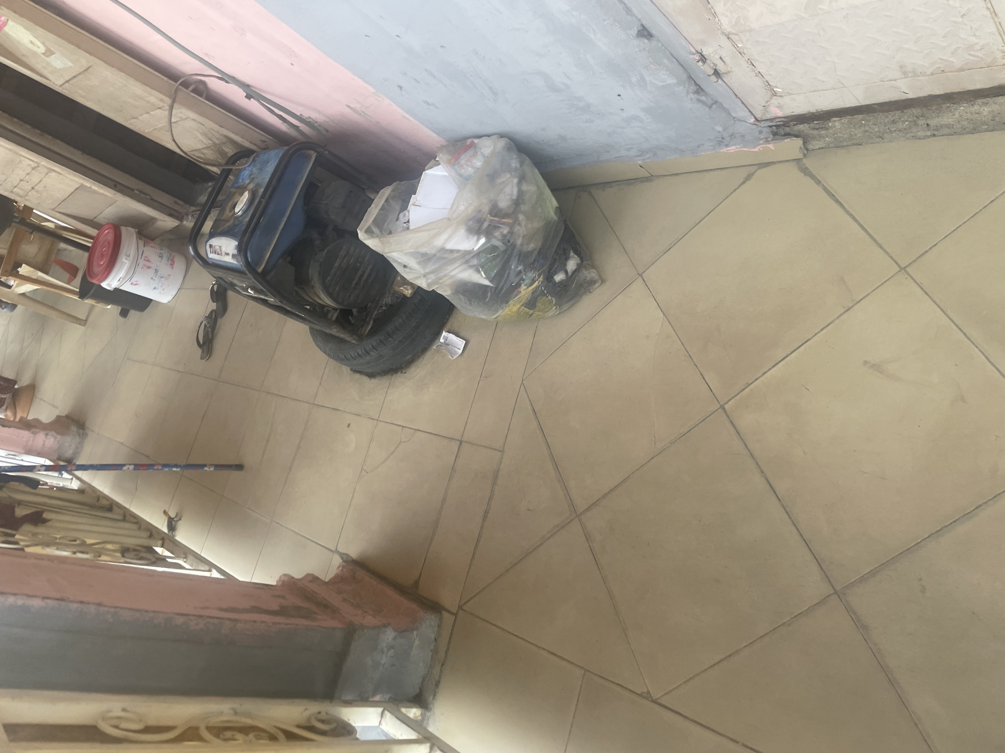 The image depicts an outdoor scene on a tiled floor, likely in Jalingo, Nigeria. In the foreground, a clear plastic bag filled with trash sits next to a blue generator with a black metal frame. A spare tire is positioned behind the generator. To the left, a partial view of a decorative pillar and railing is visible. In the background, a white bucket with a red lid and a black trash can are present, along with some wooden structures that suggest a shop or dwelling. A small lizard is visible on the tiled floor near the railing. The wall to the right appears to be painted in light pink and grey. The overall impression is of a somewhat cluttered outdoor space, possibly a residential or commercial area.