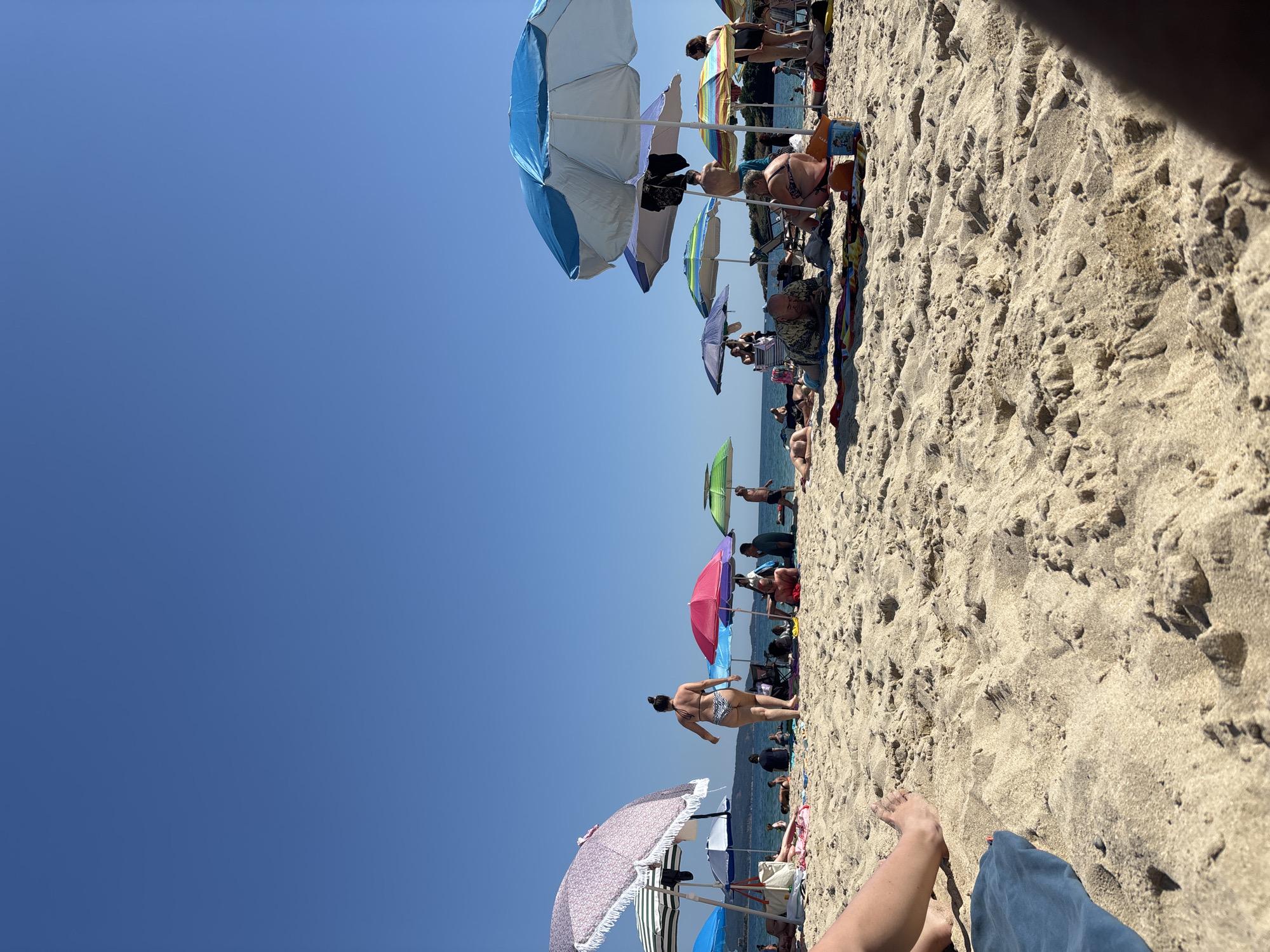 The image depicts a crowded beach in Kiten, Bulgaria, under a clear blue sky. The foreground is dominated by sand dunes and a person's leg and foot resting on a blue towel. Several beach umbrellas are spread across the sand, providing shade for beachgoers. People are seen in various states of relaxation, some lying on towels, others sitting or standing. The sea is visible in the background, with a distant coastline. The overall scene captures a typical summer day at a popular beach resort.