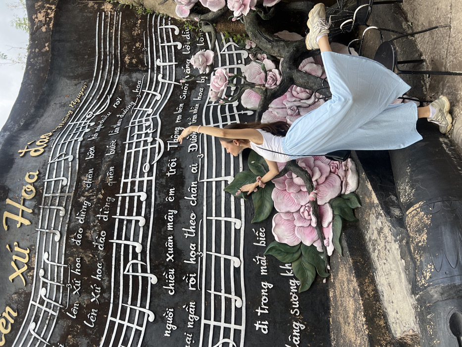 The image captures a young adult female posing in an outdoor setting at Da Lat Wonderland, Da Lat, Vietnam, in front of a large, dark-colored relief sculpture. The sculpture features white musical staves, notes, and Vietnamese text in white and gold lettering, including "Lên Xứ Hoa Đào" (To the Land of Cherry Blossoms) prominently at the top, and "Hoàng Nguyên." Intricately sculpted large pink and light green flowers with leaves and branches are integrated into the lower and right portions of the relief.
The female subject, positioned right of center, wears a white tank top, light blue wide-leg culottes, white socks, and light-colored chunky sneakers. A black shoulder bag hangs on her left side, and a pink scrunchie is on her right wrist. She is captured mid-pose, with her left foot on the ground, her right leg extended backward and upward, and her right arm raised high above her head. Her left arm is bent, with her hand positioned near her head, fingers splayed. Her head is tilted back, looking upward. A dark metal chair is partially visible behind her right leg. The ground below the sculpture is dark and uneven.