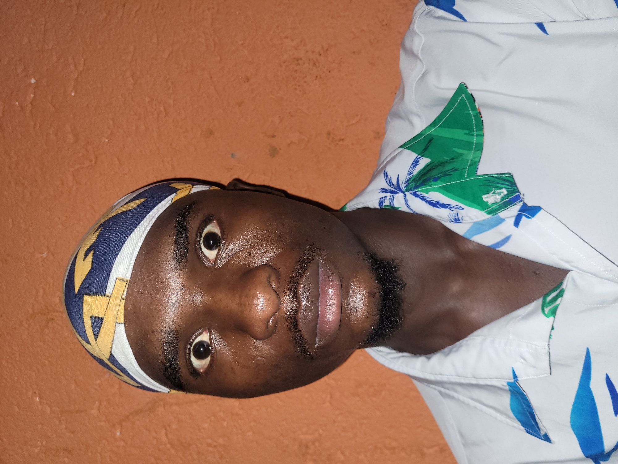 A close-up, head-and-shoulders portrait of a young Black man is presented against an orange, textured wall. The man is looking directly at the camera with wide, startled eyes. He is wearing a white shirt with a tropical print featuring palm trees and blue foliage. A bandana, patterned with blue, yellow, and white geometric shapes, is tied around his head. He has a thin mustache and a small goatee. The lighting suggests an indoor setting, possibly a home in Bukuru, Nigeria, given the man's appearance and the wall's texture, which is consistent with local construction materials.
