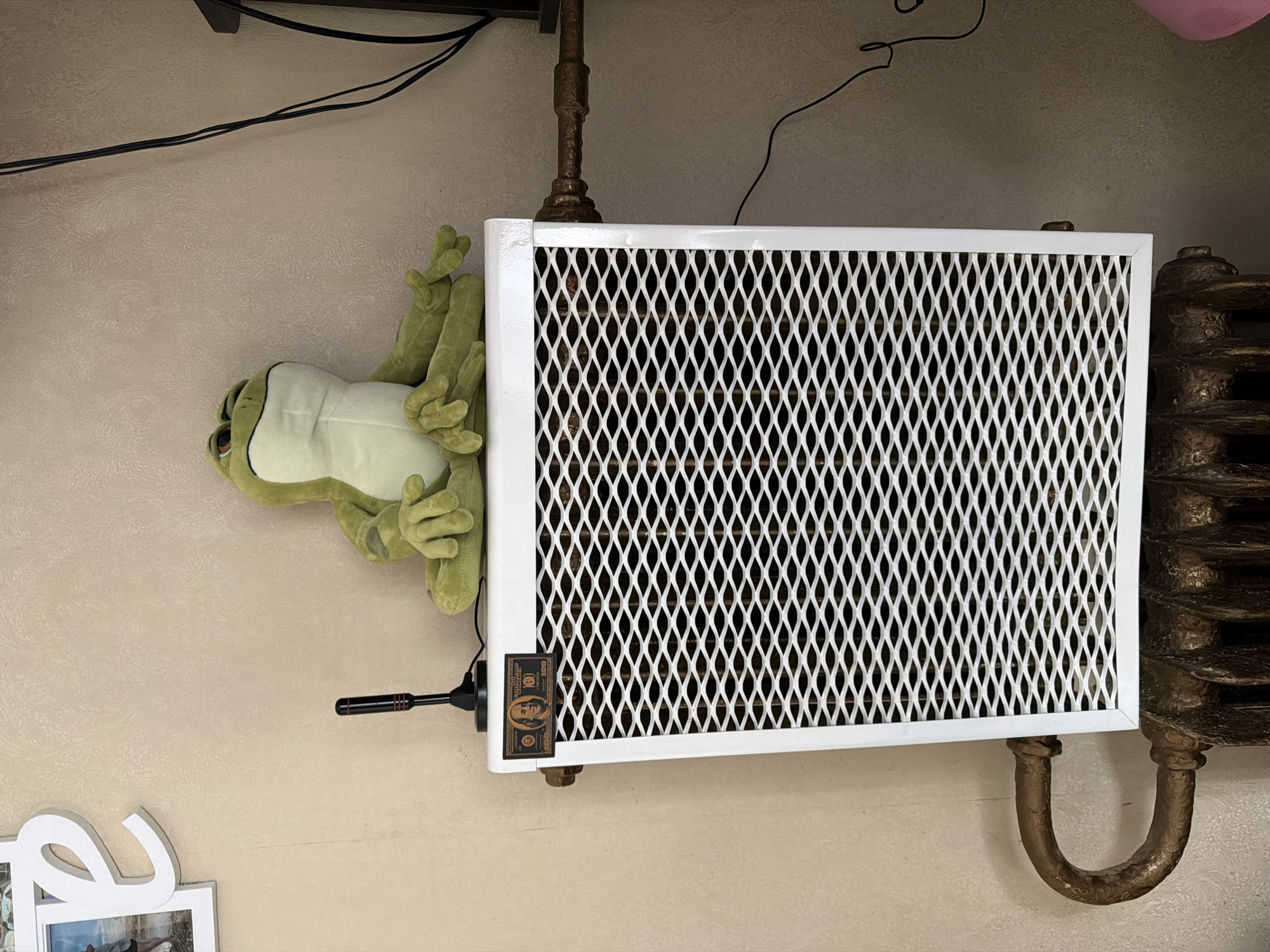 The image shows a stuffed green frog sitting on top of a white radiator cover. The radiator cover has a diamond-patterned metal grille and a decorative label resembling a US $100 bill. A small black antenna is attached to the top of the radiator cover. The radiator itself is old-fashioned, made of dark metal with a U-shaped pipe connecting it to the wall. The background is a textured beige wall. To the left of the radiator, a white decorative letter "e" is partially visible, and a framed picture is also present. To the right of the radiator, black electrical wires are seen running along the wall, and a dark object, possibly furniture or a television, is in the background. The scene appears to be indoors, in a domestic setting. The location is not specified and cannot be determined from the image.