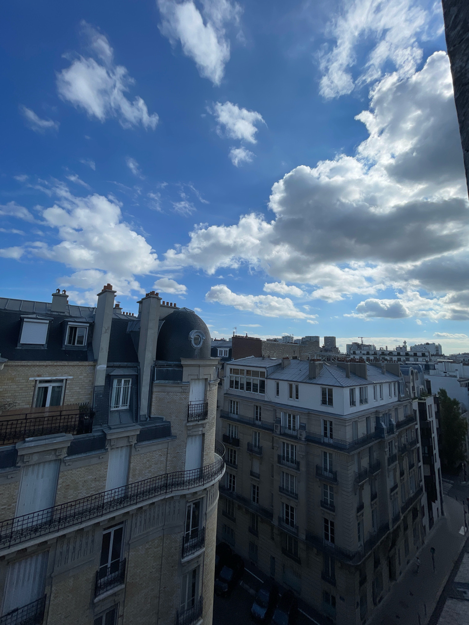 The image displays a view of Parisian rooftops under a dynamic sky with cumulus clouds. The architecture suggests an older, well-established urban district, likely within the central arrondissements of Paris, characterized by Haussmannian-style buildings with mansard roofs, dormer windows, and ornate balconies. The perspective is from a higher vantage point, looking down onto the cityscape. Prominent features include numerous chimneys, curved balconies with wrought-iron railings, and the distinctive grey zinc roofing common in Paris. The sky is a vibrant blue, interspersed with fluffy white clouds, indicating fair weather, possibly late afternoon or early morning given the lighting. No people or vehicles are clearly visible in the frame, focusing the scene entirely on the architectural elements and the natural sky.