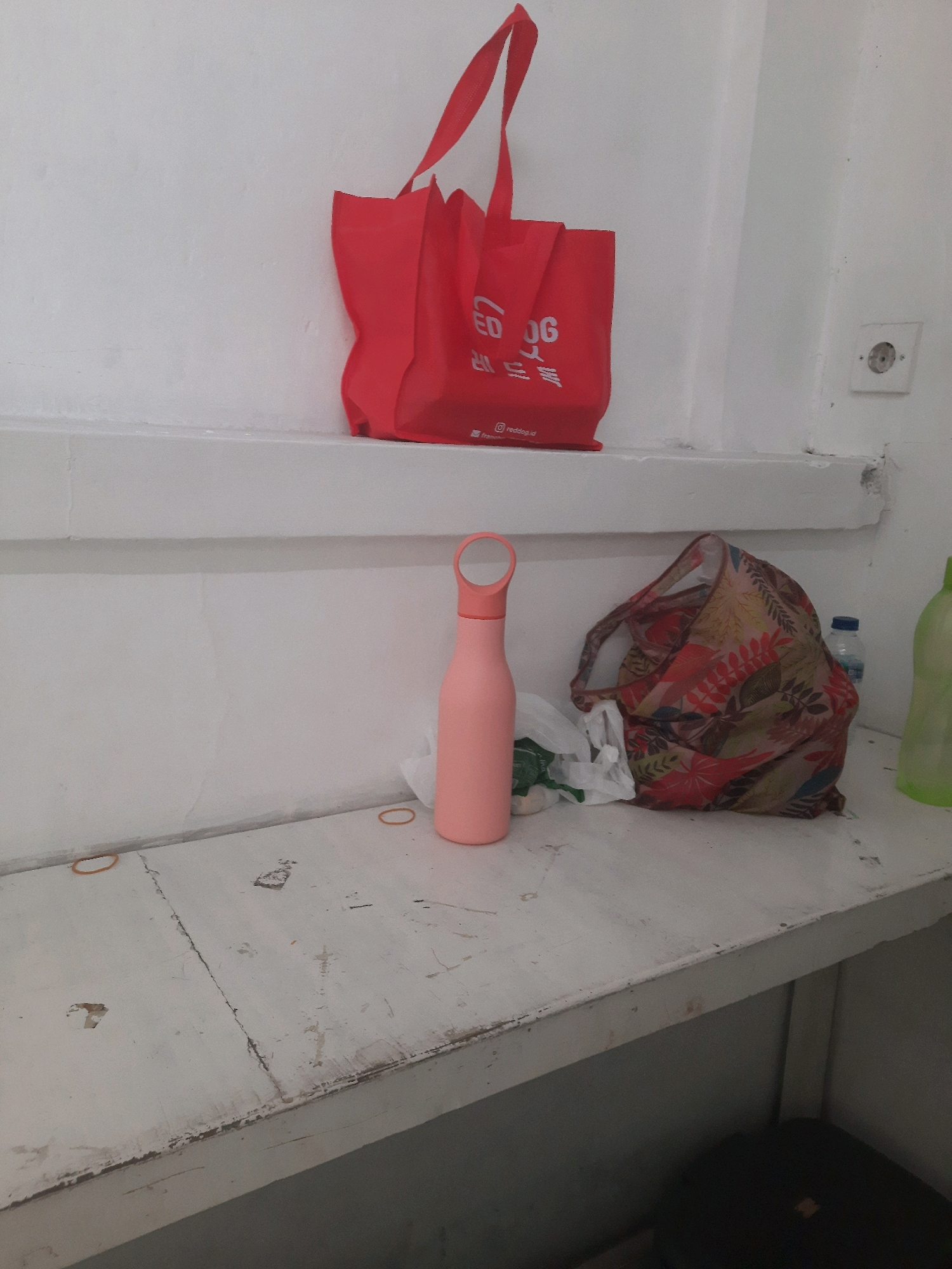 A close-up, indoor, eye-level medium shot captures a white shelf above a white wooden table in what appears to be a simple room in Padang, Indonesia. On the shelf, a bright red reusable shopping bag with white lettering is prominently displayed. To the right of the bag, a small white electrical outlet with a circular port is visible on the white wall.
On the table below, a pink reusable water bottle stands upright on the left. Next to it, a plastic bag contains what appear to be white items. Further to the right, a patterned reusable bag with a floral design is partially open. Behind these items, a clear plastic bottle of water is situated. On the far right edge of the table, a portion of a green plastic bottle is visible. The surface of the wooden table is worn and shows signs of age and use, with cracks and peeling paint. Two thin brown rubber bands are also visible on the table surface, one near the water bottle and another further to the right. In the lower right corner, a dark object, possibly a seat or stool, is partially visible. The overall scene suggests a practical, everyday setting.