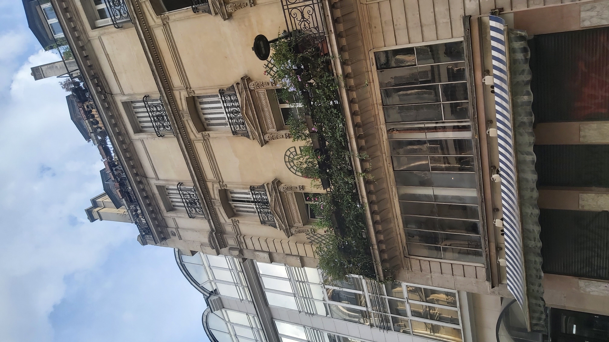 This is a low-angle, eye-level exterior shot of a Parisian building. The building is made of pale stone with ornate architectural details, including decorative window surrounds, balconies with wrought iron railings, and a Mansard roofline with dormer windows. There are multiple floors visible, with windows featuring closed shutters. A prominent balcony on an upper floor is heavily laden with lush green plants and flowers. Adjacent to this building is a more modern structure with a large, curved glass facade, reflecting the sky and the older building. The foreground shows the lower part of the older building, which includes large windows and a striped awning above what appears to be storefront shutters. The sky is partly cloudy. The scene is located in Paris, France.