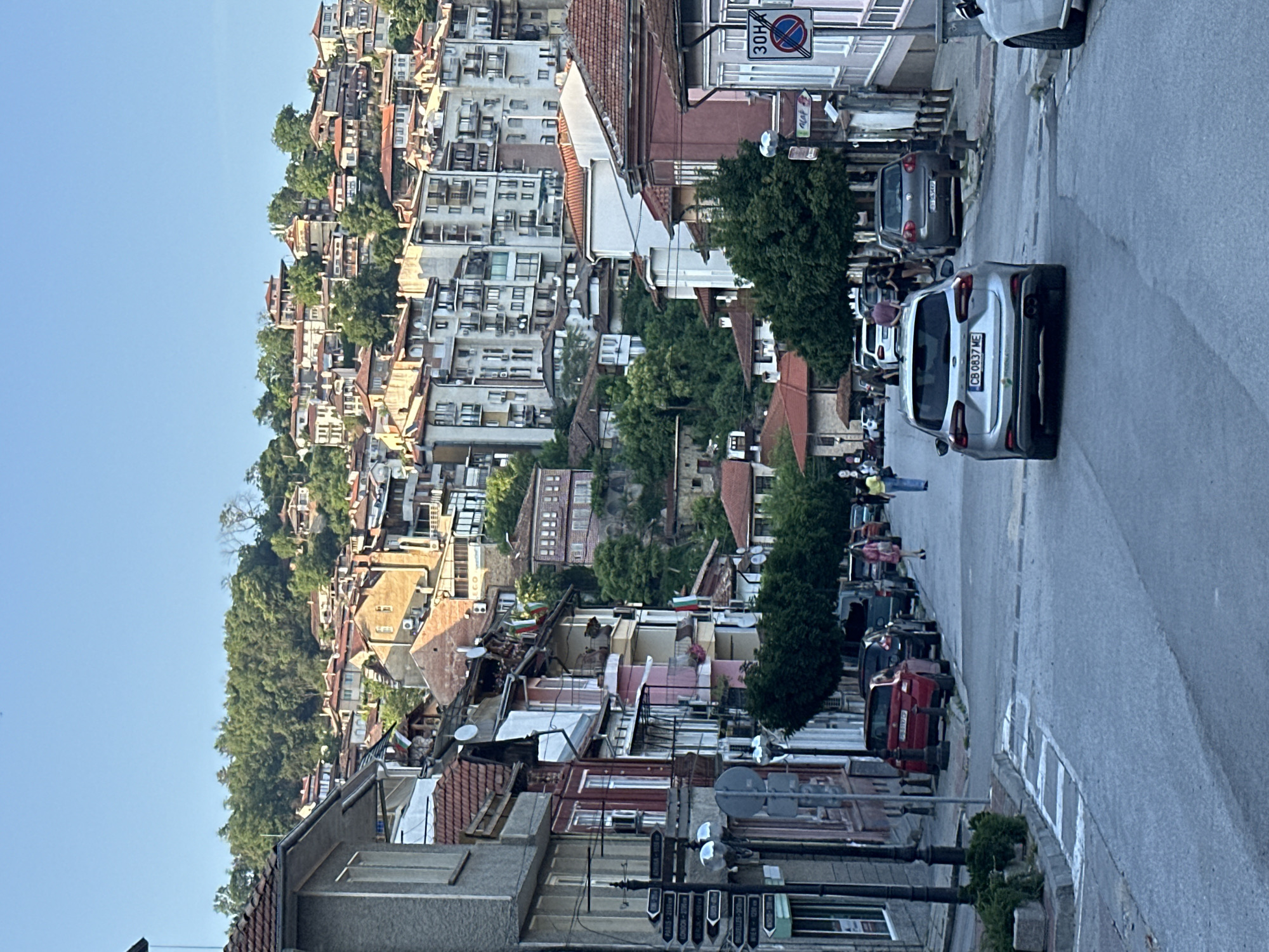 The image presents an ascending street in Veliko Tarnovo, Bulgaria, terminating at a hillside densely populated with buildings. The sky above is clear and blue.
In the foreground, an asphalt road occupies the center, showing evidence of pedestrian crossing markings. A silver Ford Focus (Bulgarian license plate CB 0837 ME) is centrally positioned, facing away from the viewer. Behind it, a dark grey Volkswagen Golf (Bulgarian license plate BT 5344 KP) is partially visible, parked on the right side of the road with other vehicles. A red car (Bulgarian license plate PB 3197 CP) is parked further back on the left.
Along the left side of the street, a black street sign pole features multiple directional arrows with white text, indicating distances to international cities: Paris (2190 km), London (2300 km), Berlin (1550 km), Athens (1280 km), and Vienna (1200 km). Buildings of varying architectural styles line this side, some with balconies and satellite dishes.
On the right side of the street, more multi-story buildings are present. A circular traffic sign with a red border and diagonal line contains "30H," indicating a time-limited restriction, likely for parking or stopping. Bulgarian flags are visible hanging from balconies of several structures. Small, manicured trees line both sides of the street, and several pedestrians are walking on the sidewalks and in the road.
The midground and background feature a steep, green hillside extensively developed with residential buildings. Numerous multi-story structures, predominantly with red-tiled roofs, are tightly packed, showcasing a mix of older and more modern architectural designs. Dense green foliage and trees are interspersed among the buildings, particularly on the higher slopes.