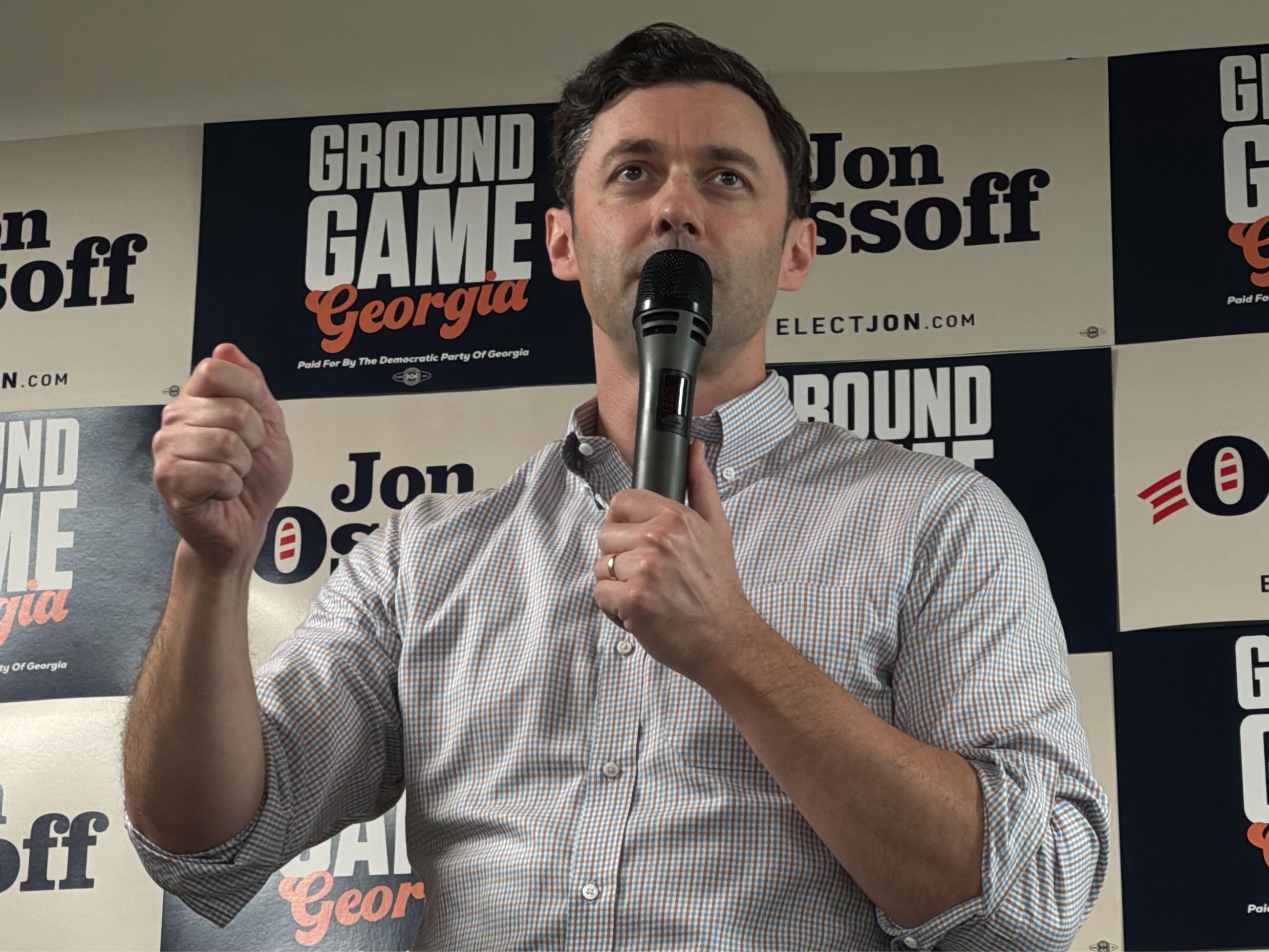 A man speaks into a microphone, standing in front of a backdrop promoting Jon Ossoff's campaign. The backdrop includes campaign slogans such as "Ground Game Georgia" and "Elect Jon". The man wears a checkered button-down shirt and a ring on his left hand. He is holding the microphone with one hand, and his other hand is raised in a fist. The location is the city of Decatur, United States.