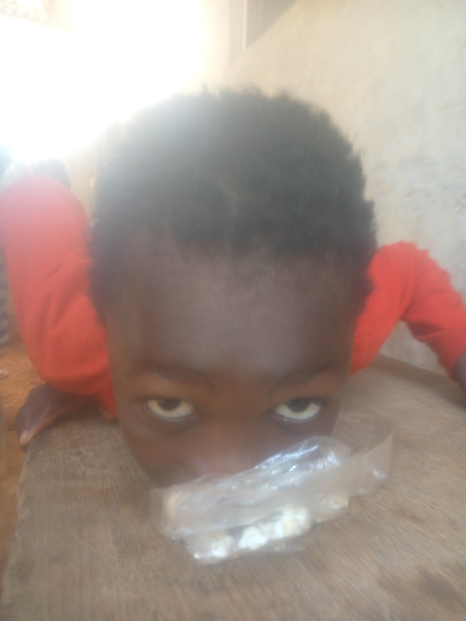 A close-up, low-angle photograph captures a young child with dark skin and short, dark, curly hair. The child is wearing a red long-sleeved shirt and is positioned on a wooden surface, likely a table or floor, looking directly into the camera. Their head is tilted forward, bringing their eyes into prominent focus, displaying a direct and slightly intense gaze.
A small, clear plastic bag containing several white pieces of food, possibly popcorn or similar snacks, is positioned in front of the child's mouth, partially obscuring it. The child's hands are visible resting on the wooden surface on either side of their head. The background is heavily overexposed and bright white, suggesting a strong light source, possibly from a window or outdoors, and makes it difficult to discern specific details of the setting beyond a general sense of an indoor, possibly simple, environment. The image appears to be taken during the daytime. No visible text is present in the image.