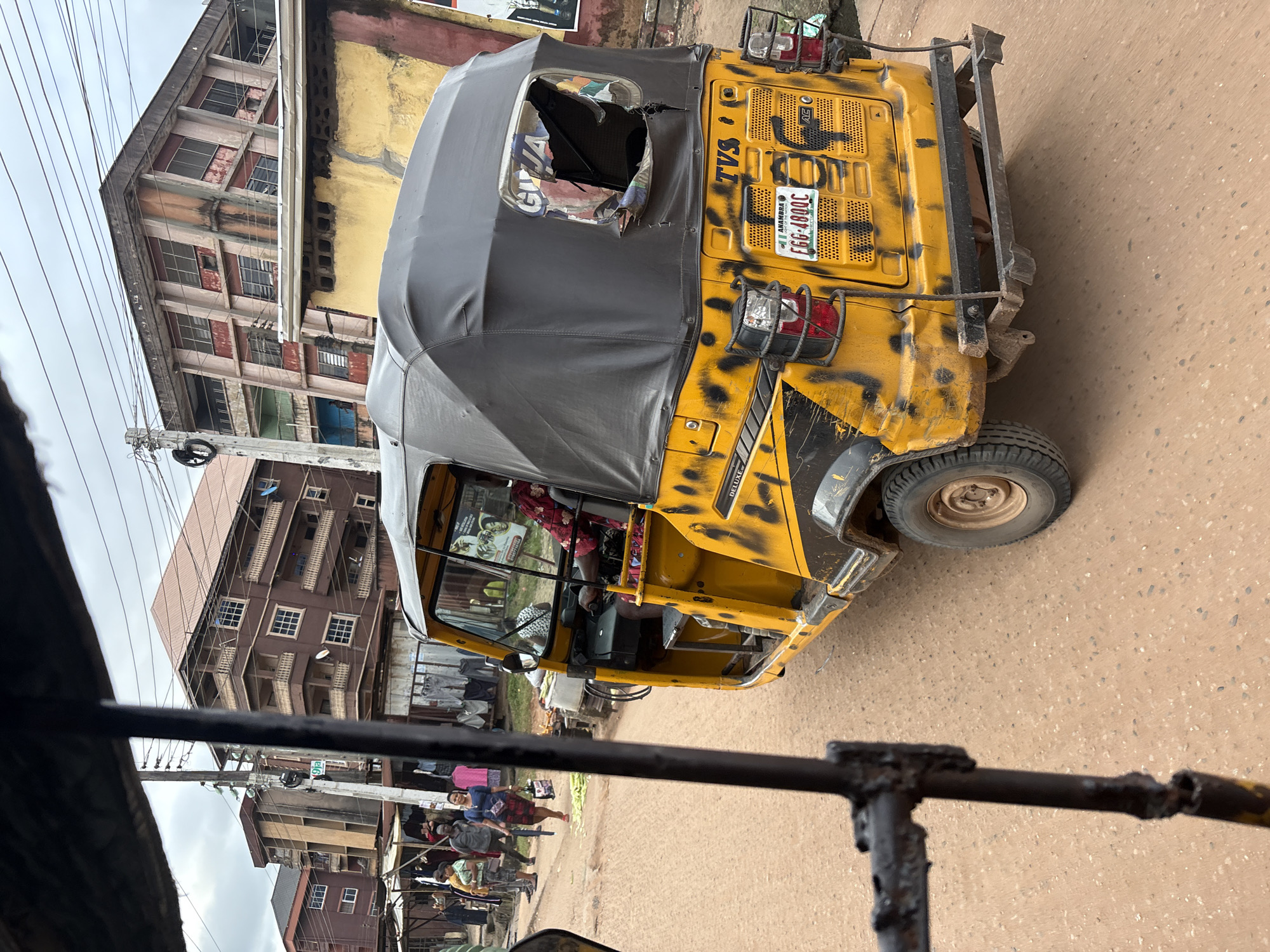 A yellow and black-patterned auto-rickshaw is parked on a dusty, unpaved road in Nkpor, Nigeria. The auto-rickshaw has a black fabric roof and a broken rear window. The license plate reads "EGG 4800C" and is from Anambra State. In the background, there are several multi-story buildings with balconies and open windows. A power pole with wires runs along the street. Several people are walking in the background, some carrying bags. Clothing racks are set up in front of one of the buildings, displaying various garments. The scene appears to be a busy street market or commercial area.