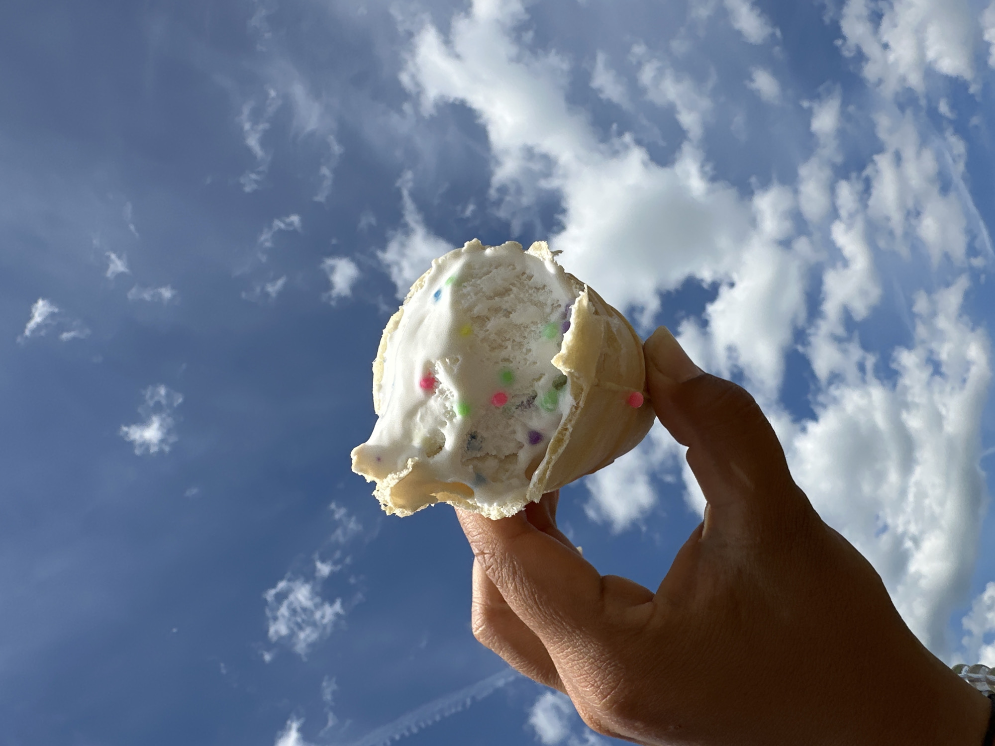 A hand with brown skin holds a cone with white ice cream. The ice cream is topped with colorful sprinkles. The background is a blue sky with white clouds. The image is taken from a low angle, looking up at the sky.