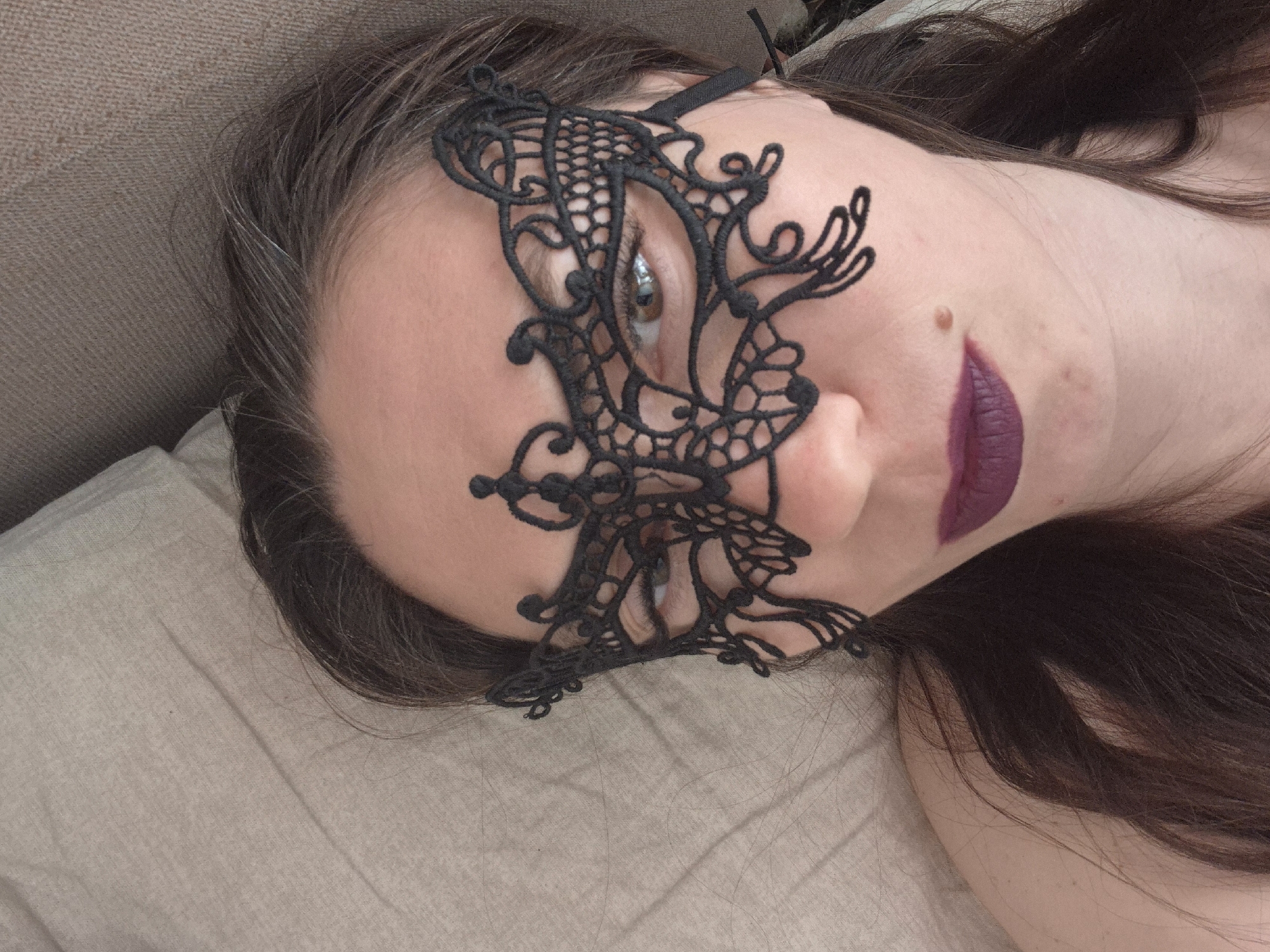A woman lies on a light beige surface, likely a bed or sofa, wearing a black lace mask that covers the upper half of her face. Her dark brown hair is spread around her head, with some gray strands visible. She has fair skin and wears dark purple lipstick. Her expression is neutral. The location is Unknown, Unknown.