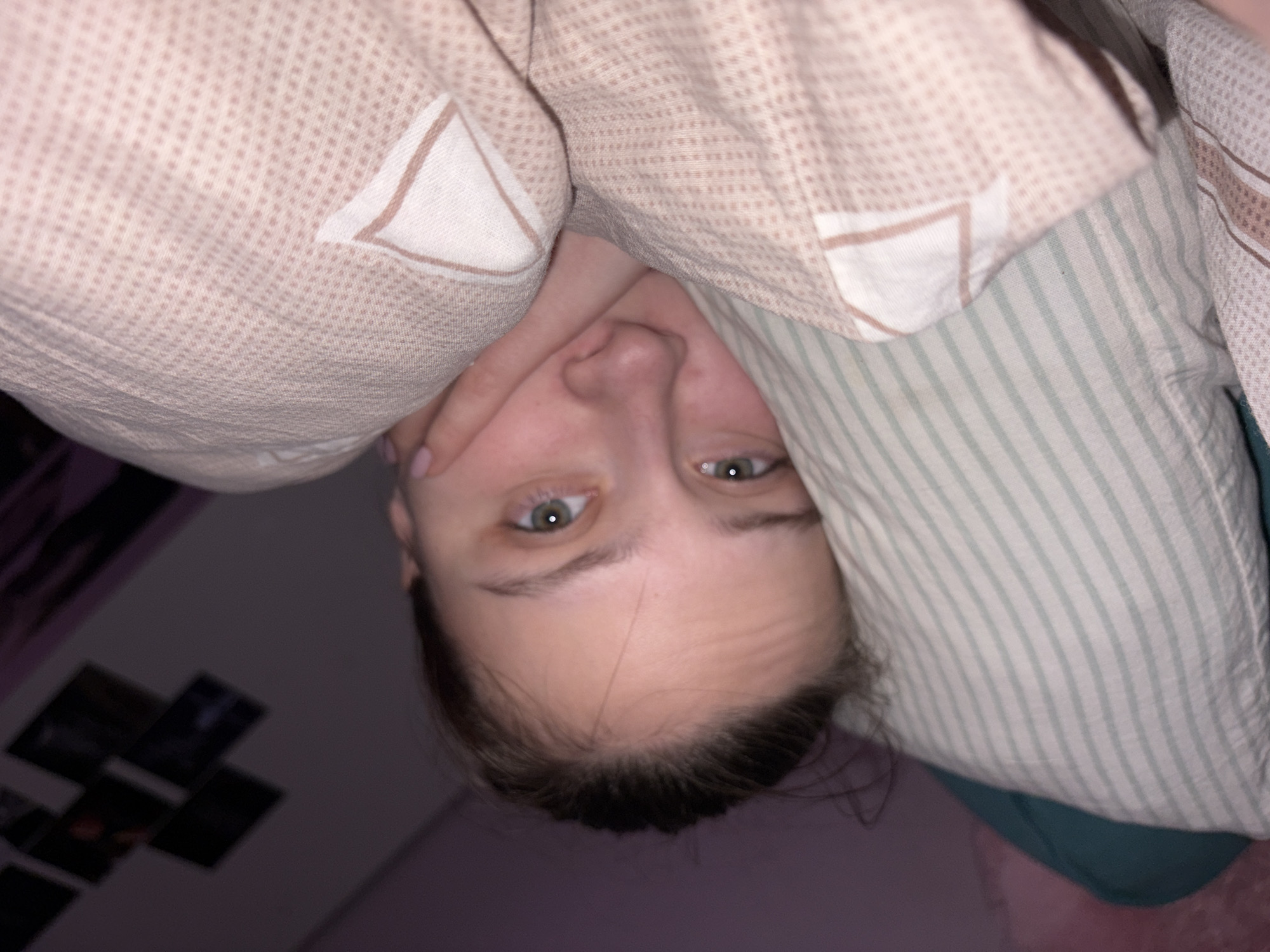 A person lies in bed under bed linens and a pillow with green and white stripes. The person's eyes are open and looking at the camera. The person's hand is visible near their face. There are blurred photographs on the wall in the background. The location is inside, presumed to be a bedroom, and the image's origin is likely Russia. No further information is discernible from the provided image.