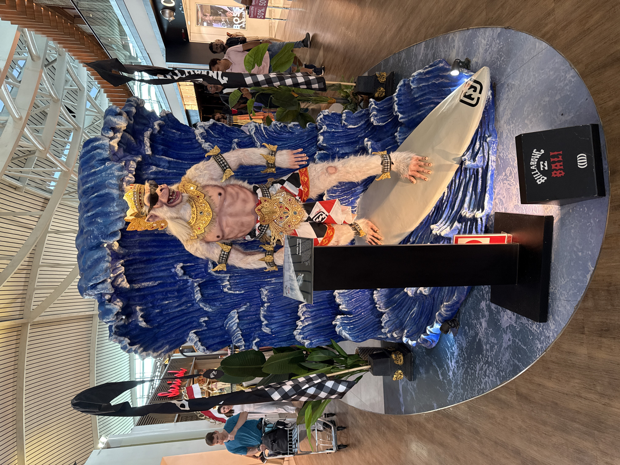 An outdoor scene depicts a large, elaborate sculpture of a monkey in a regal headdress and sunglasses, appearing to surf on a surfboard. The sculpture is set against a backdrop of giant blue waves, with black and white checkered flags and potted plants flanking it. The sculpture is situated on a circular platform, which in turn rests on a wooden floor. In the background, several people are visible walking by, some appearing to be shopping in stores with signage. The overall environment suggests a public space, possibly an airport or a shopping mall, given the lighting and architectural elements of the ceiling. The presence of a "Billabong Bali" sign near the base of the sculpture strongly suggests the location is Kuta, Indonesia, a popular tourist destination known for its surfing culture and shopping.