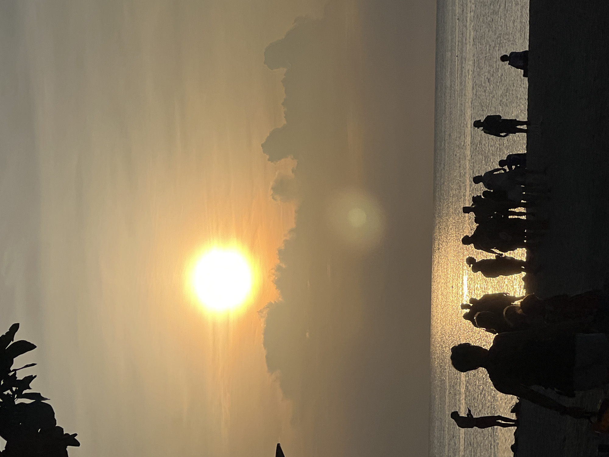 The image captures a sunset on a beach in the Ko Pha-ngan District, Thailand. Silhouettes of people are visible on the sandy shore and near the water's edge. The sun, a bright, luminous orb, is low in the sky, casting a golden glow across the clouds and the ocean surface. Some individuals are standing, some appear to be walking, and others are seated. The sea is calm, reflecting the warm hues of the sunset. The foreground is dark, with the silhouettes of trees or foliage visible at the top left, framing the scene.